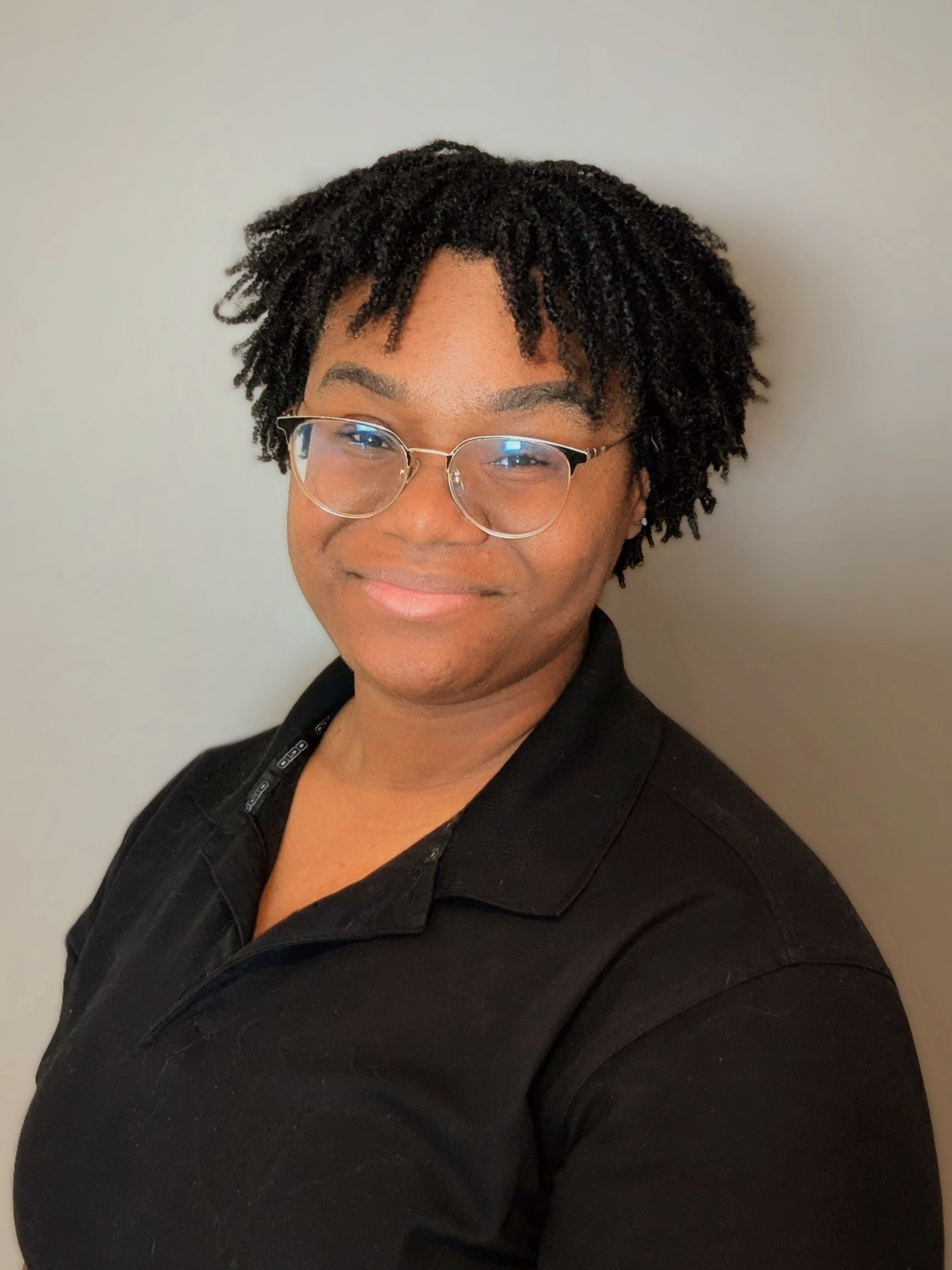 Person with glasses, short dreadlocks, smiling, wearing a black shirt, against a gray wall.