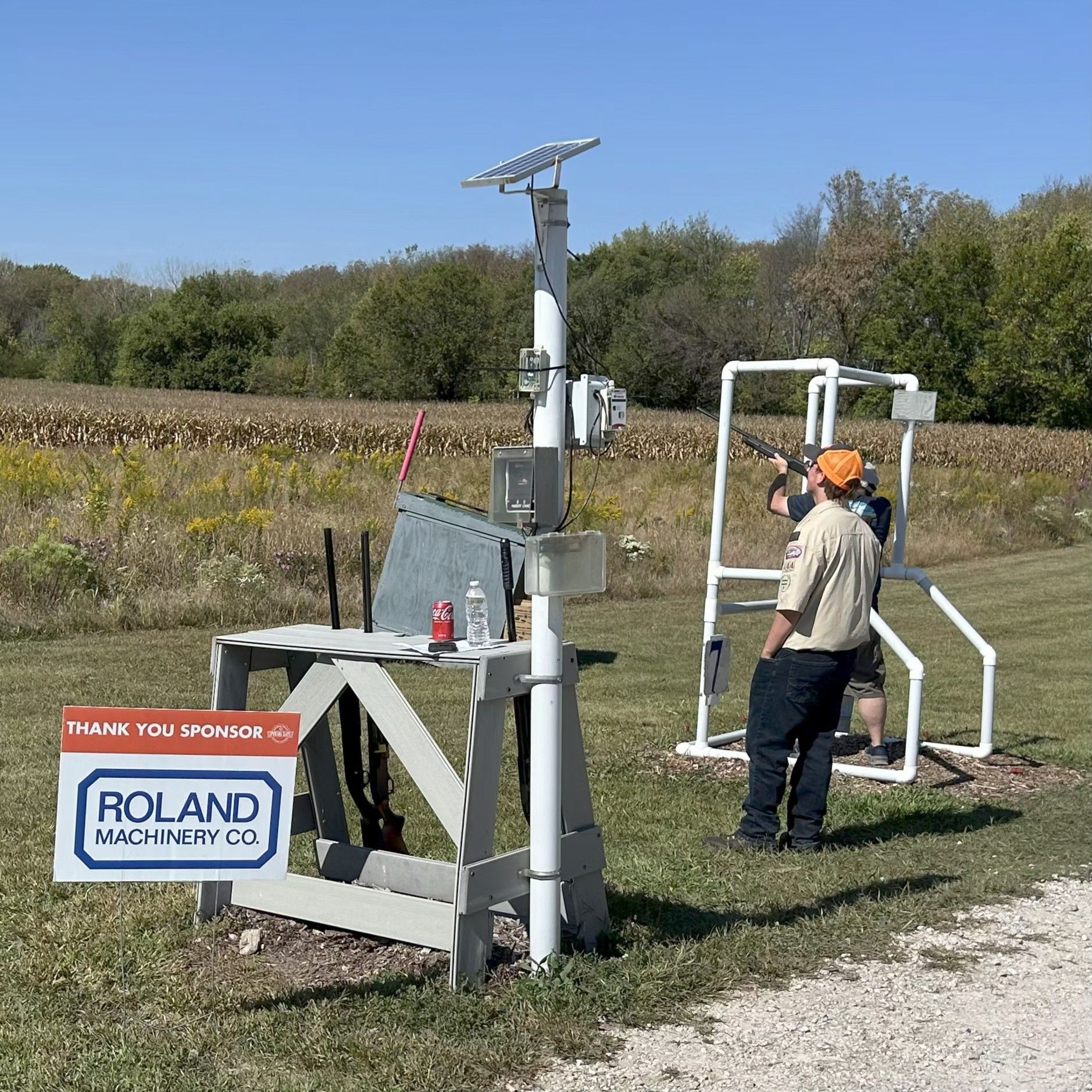 People shooting shotguns at a range; some wearing safety gear, standing on a grassy field.