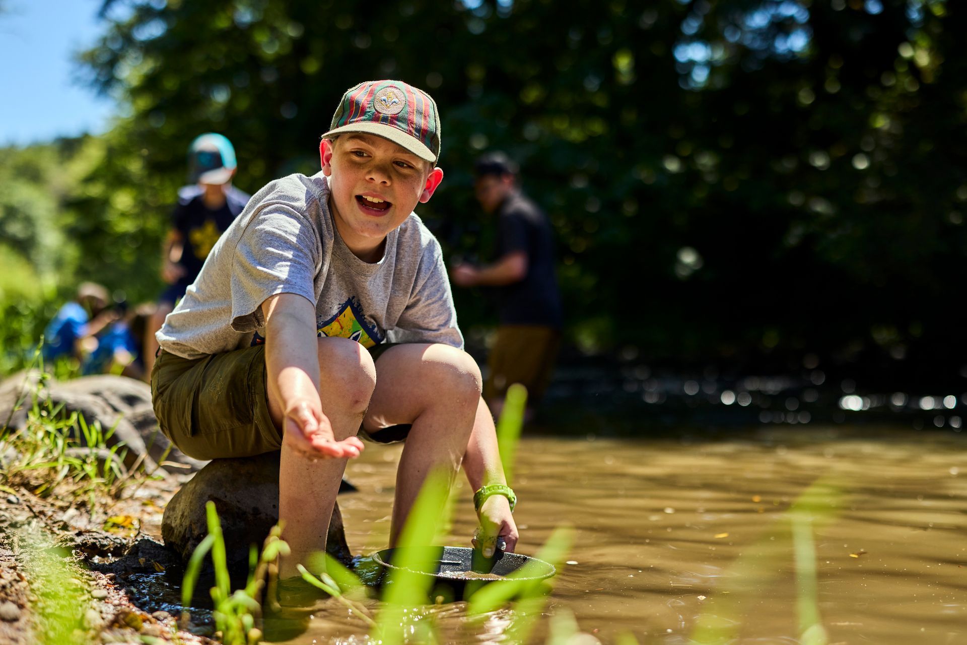 Boy in a cap sitting on a rock by a stream, smiling, reaching into water.