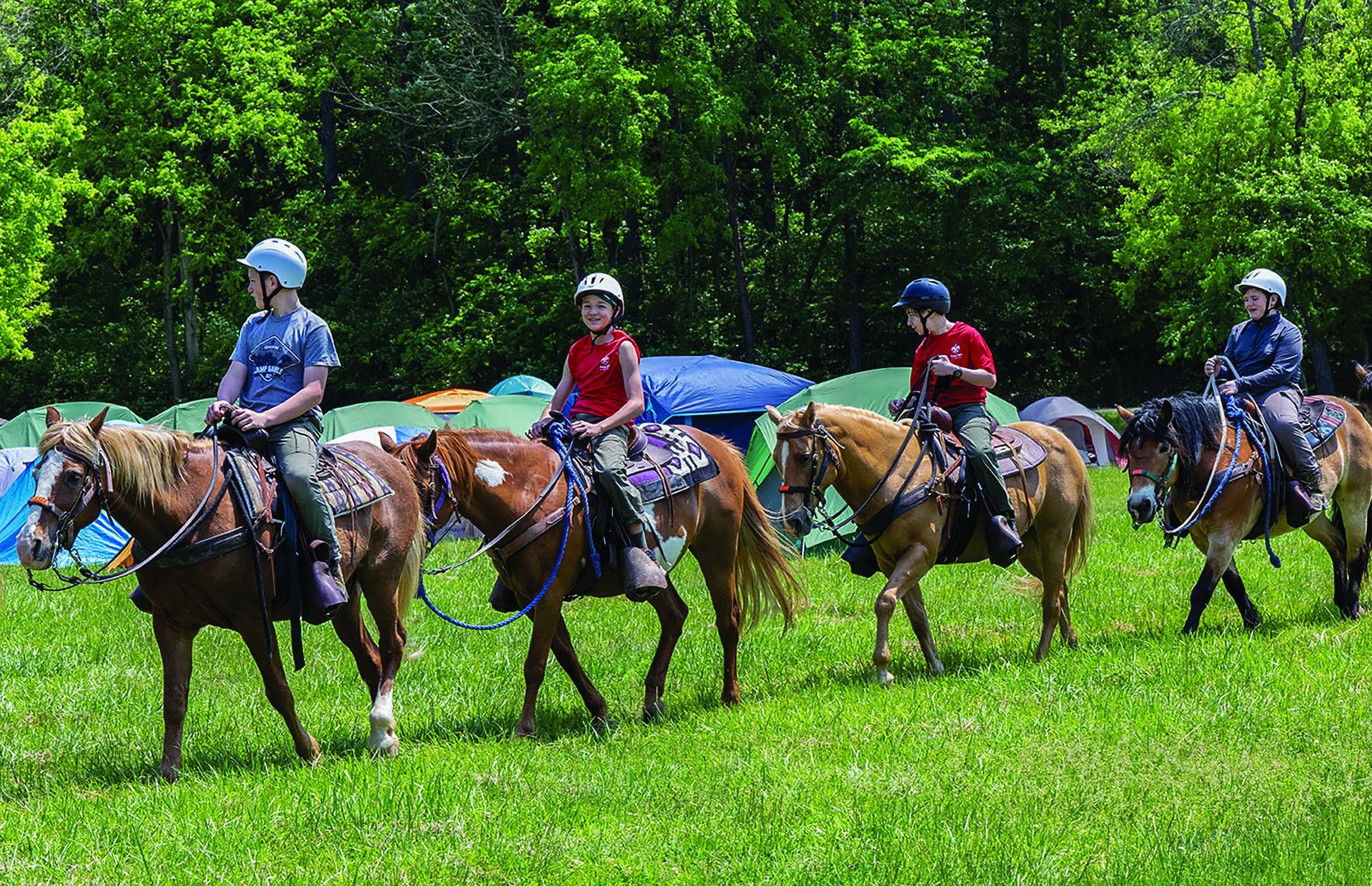 Four people on horseback ride on a grassy field in front of tents.