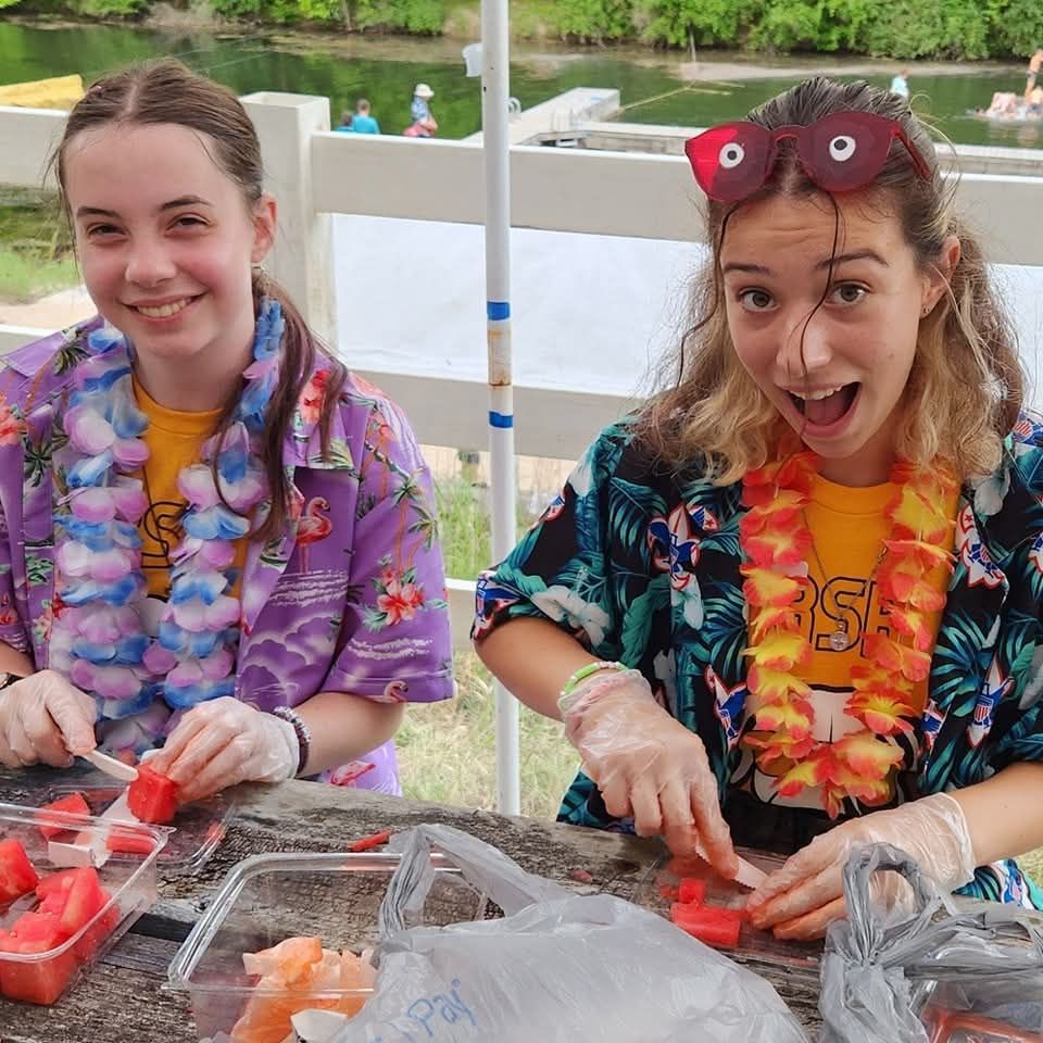 Two people in scout uniforms holding a dark tarp, with trees and sunlight in the background.