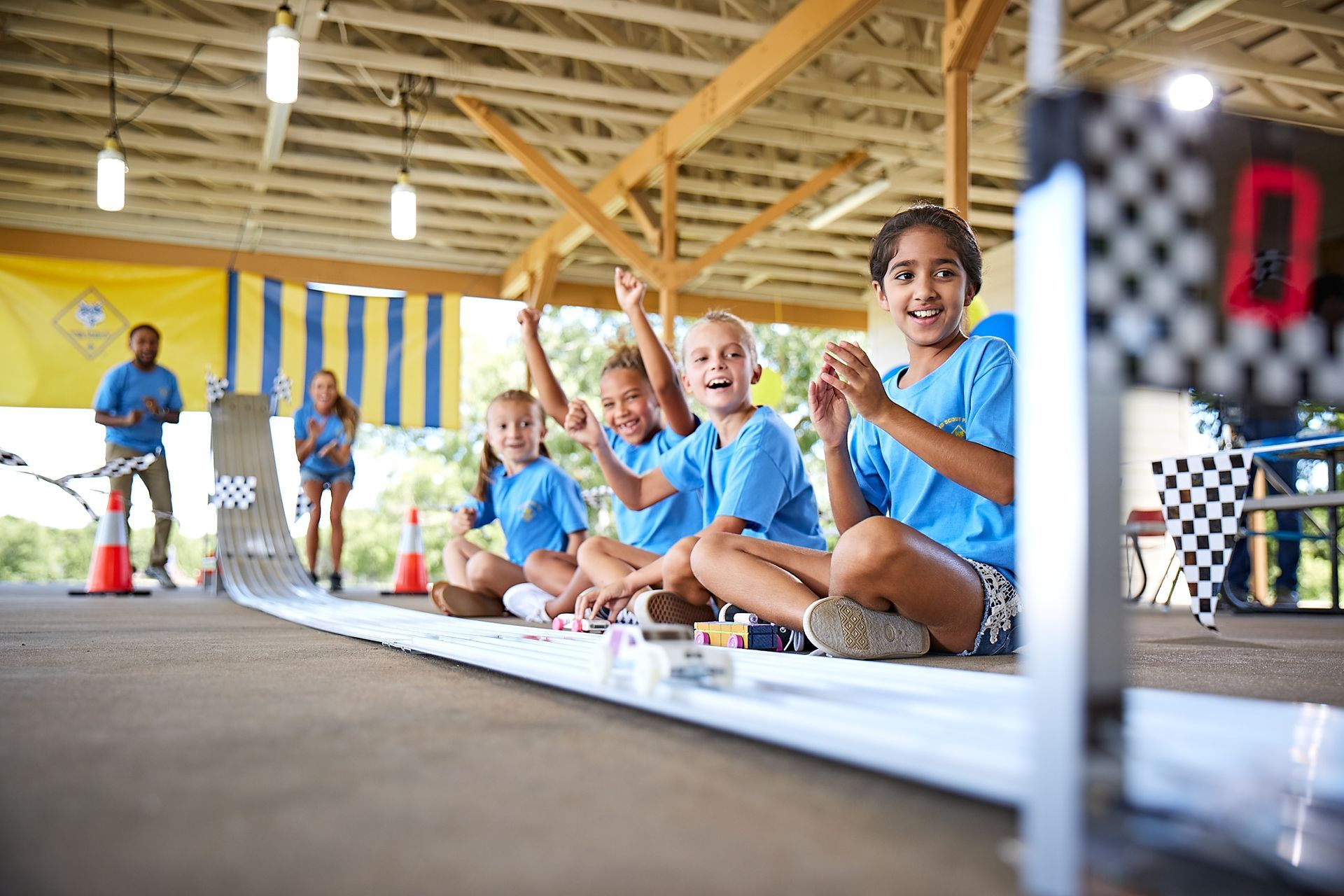 Children cheering, sitting by a model car race track, under a covered pavilion.