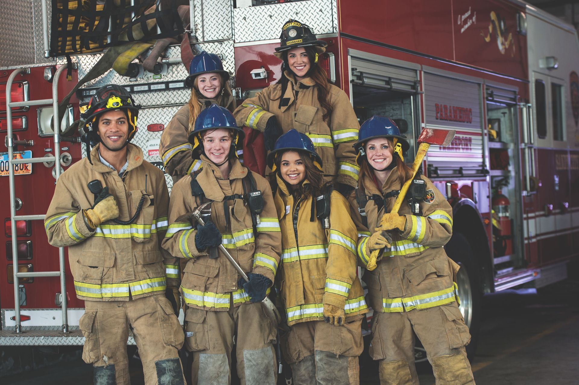 Firefighters pose in front of a fire truck. They wear tan coats and helmets, some holding tools.