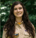Woman with long brown hair, smiling. Wearing a tan uniform outdoors.
