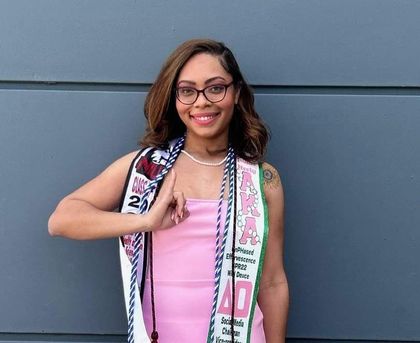 A woman wearing glasses and a pink dress is standing in front of a wall.