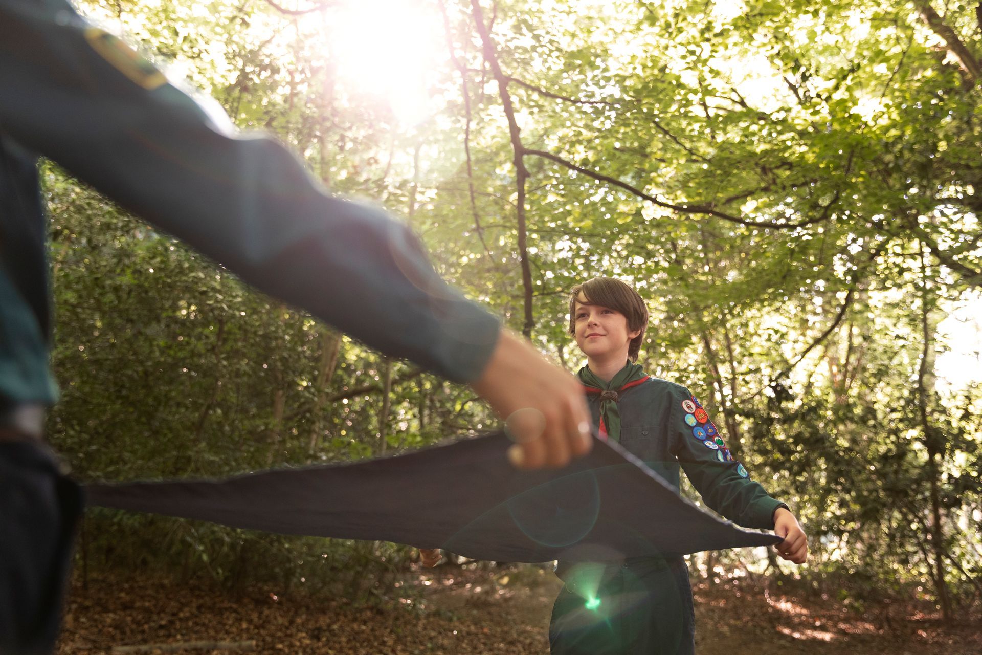 Two people in scout uniforms holding a dark tarp, with trees and sunlight in the background.