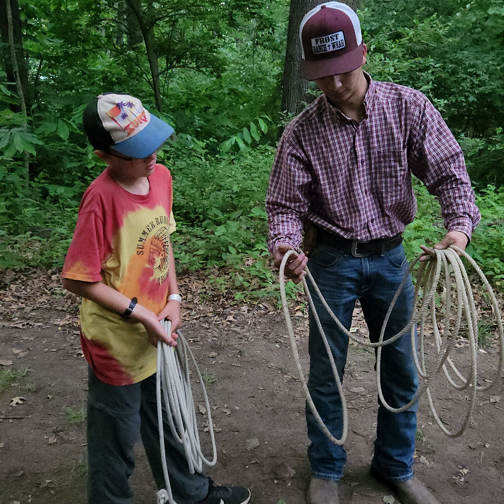 Two people in scout uniforms holding a dark tarp, with trees and sunlight in the background.