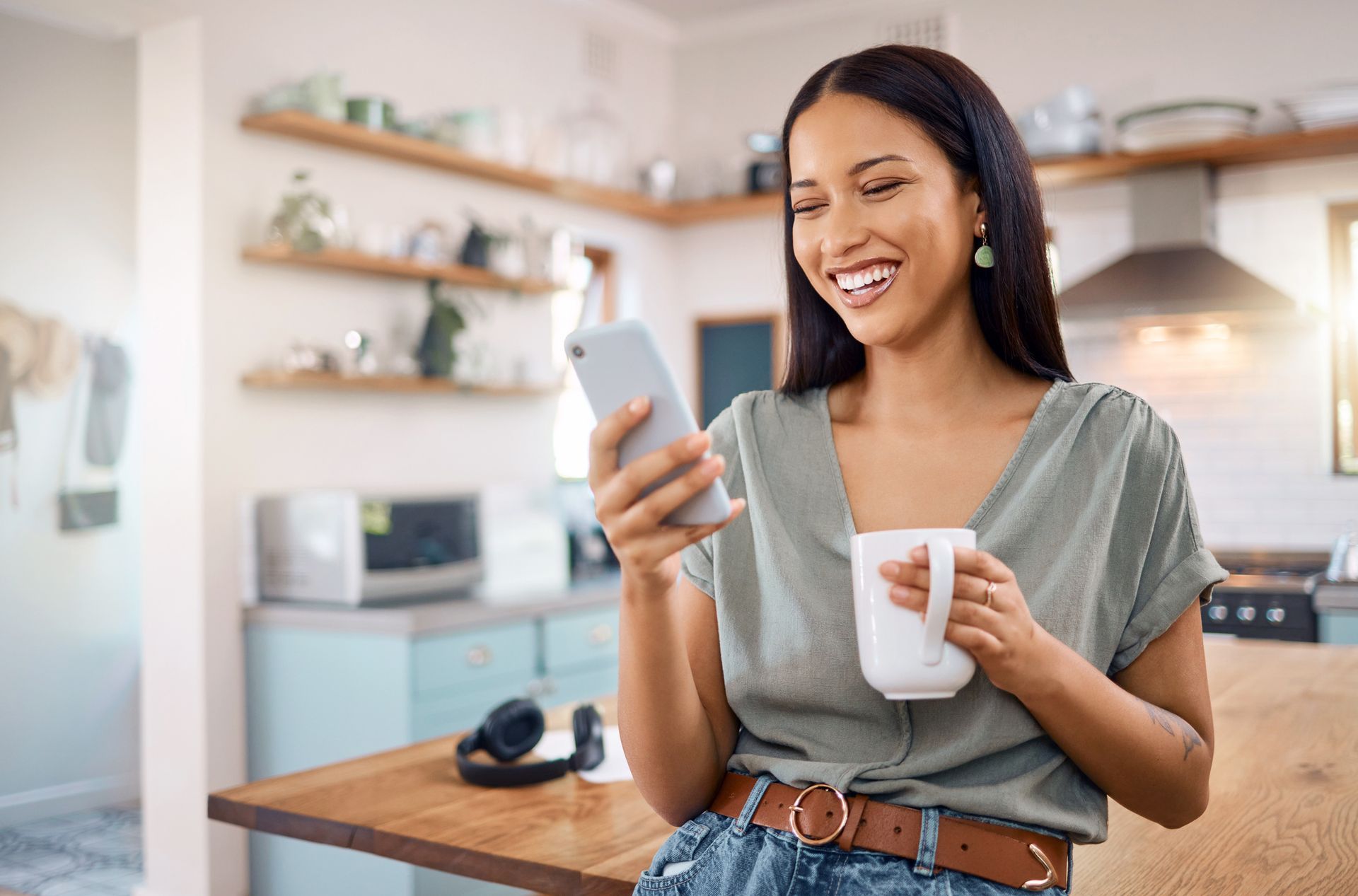 Woman in kitchen holding phone and mug, smiling.