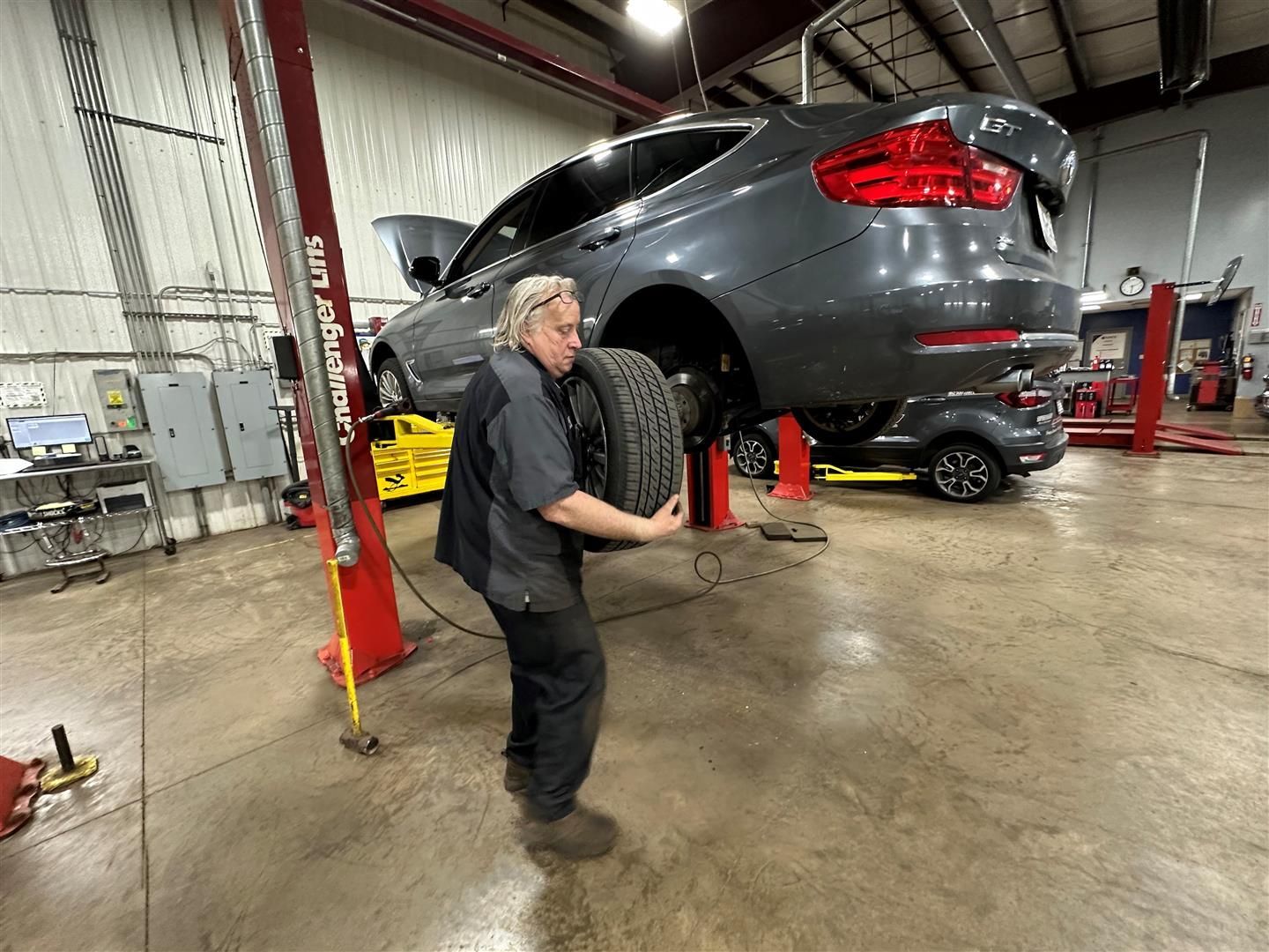 A man is holding a tire in front of a car on a lift in a garage.  | Lou's Car Care Center, Inc.