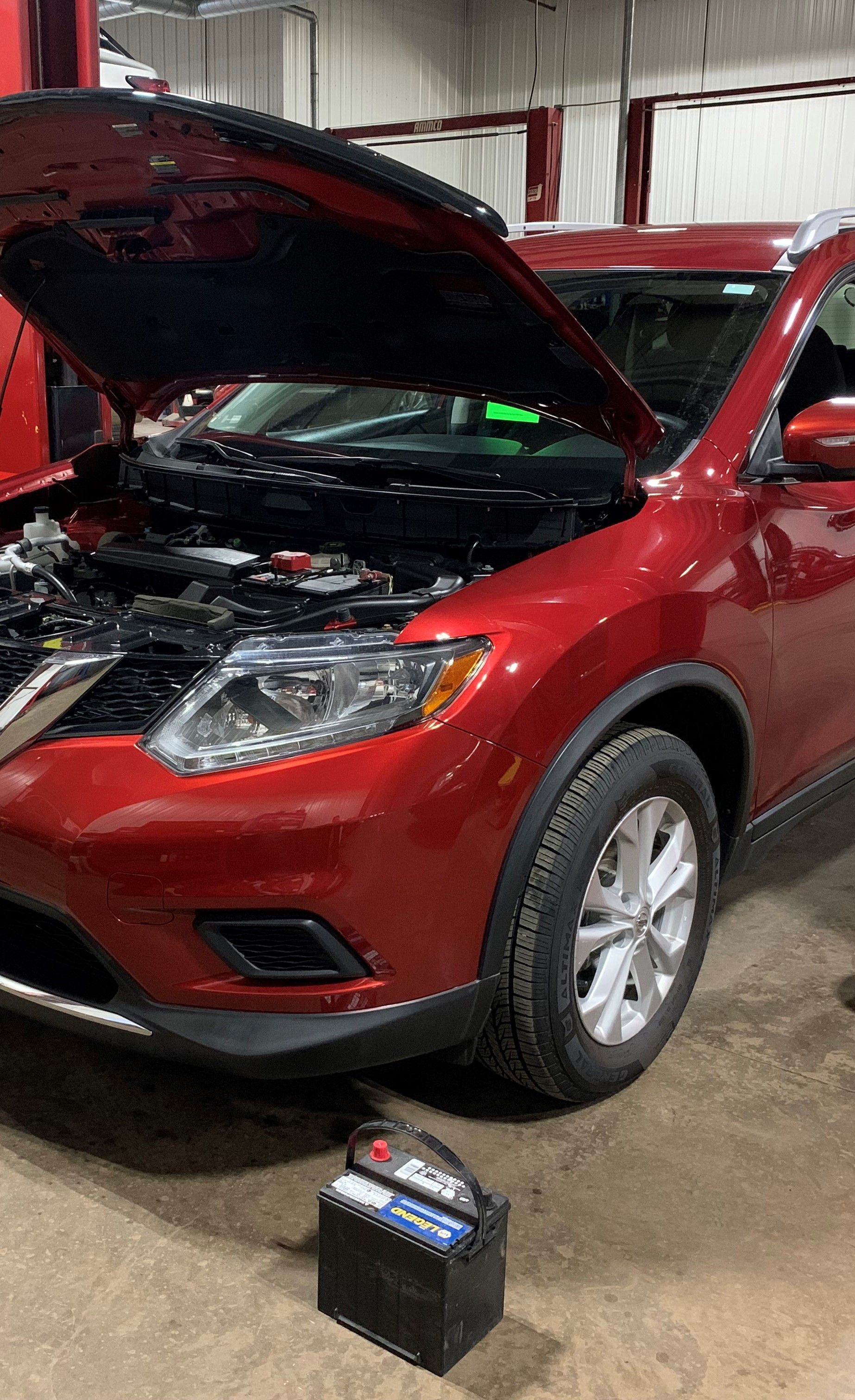 A red car with the hood open is sitting in a garage next to a battery. | Lou's Car Care Center, Inc.