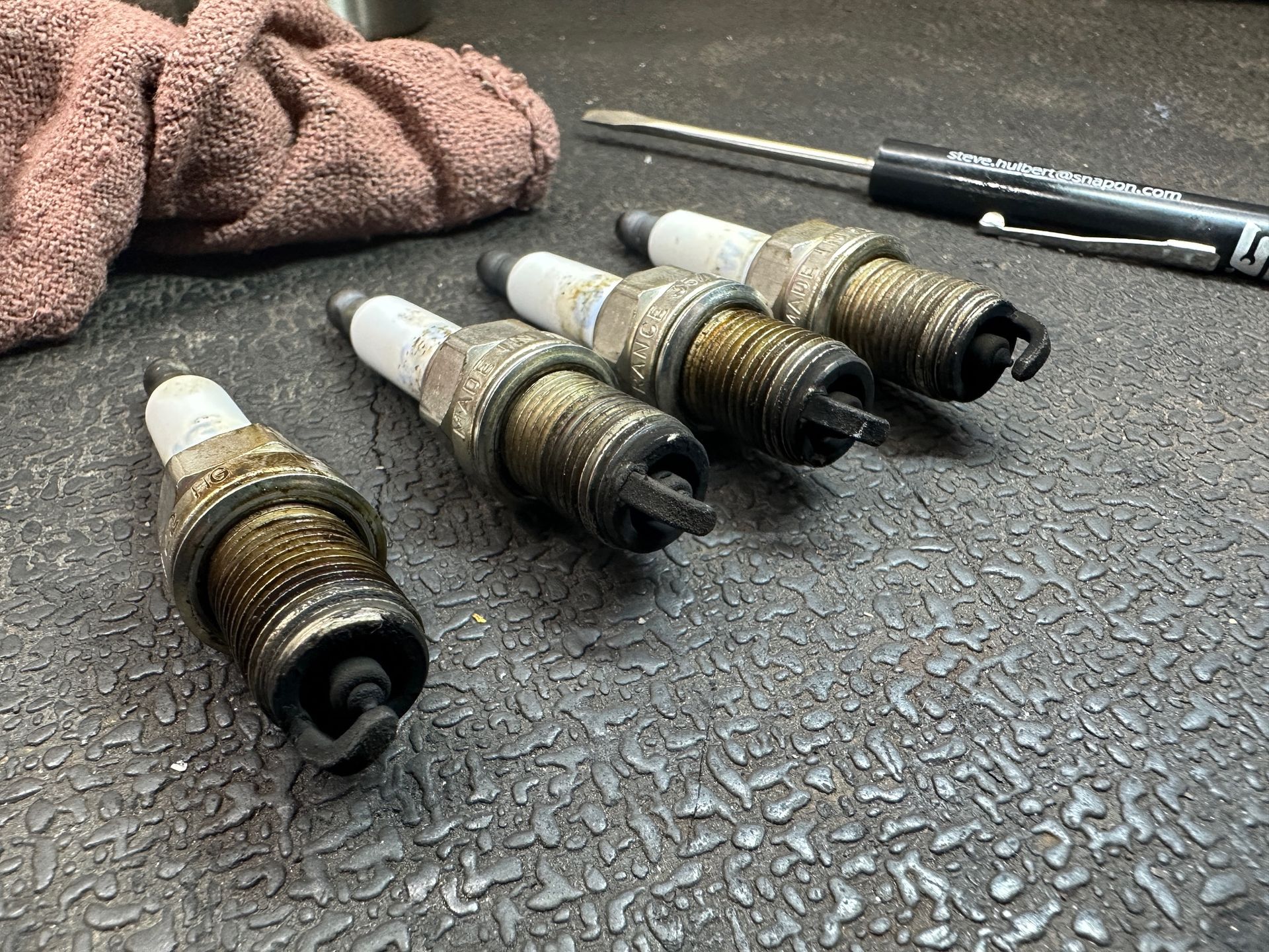 Four spark plugs are sitting on a table next to a screwdriver and a towel. | Lou's Car Care Center, Inc.