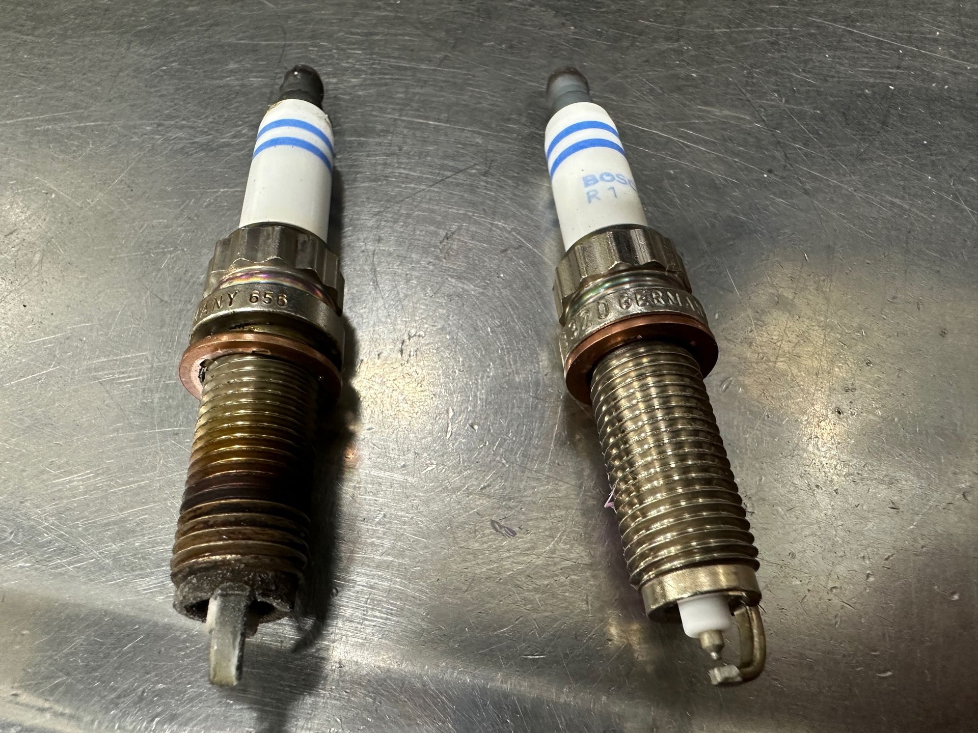 Two spark plugs are sitting next to each other on a metal surface. | Lou's Car Care Center, Inc.