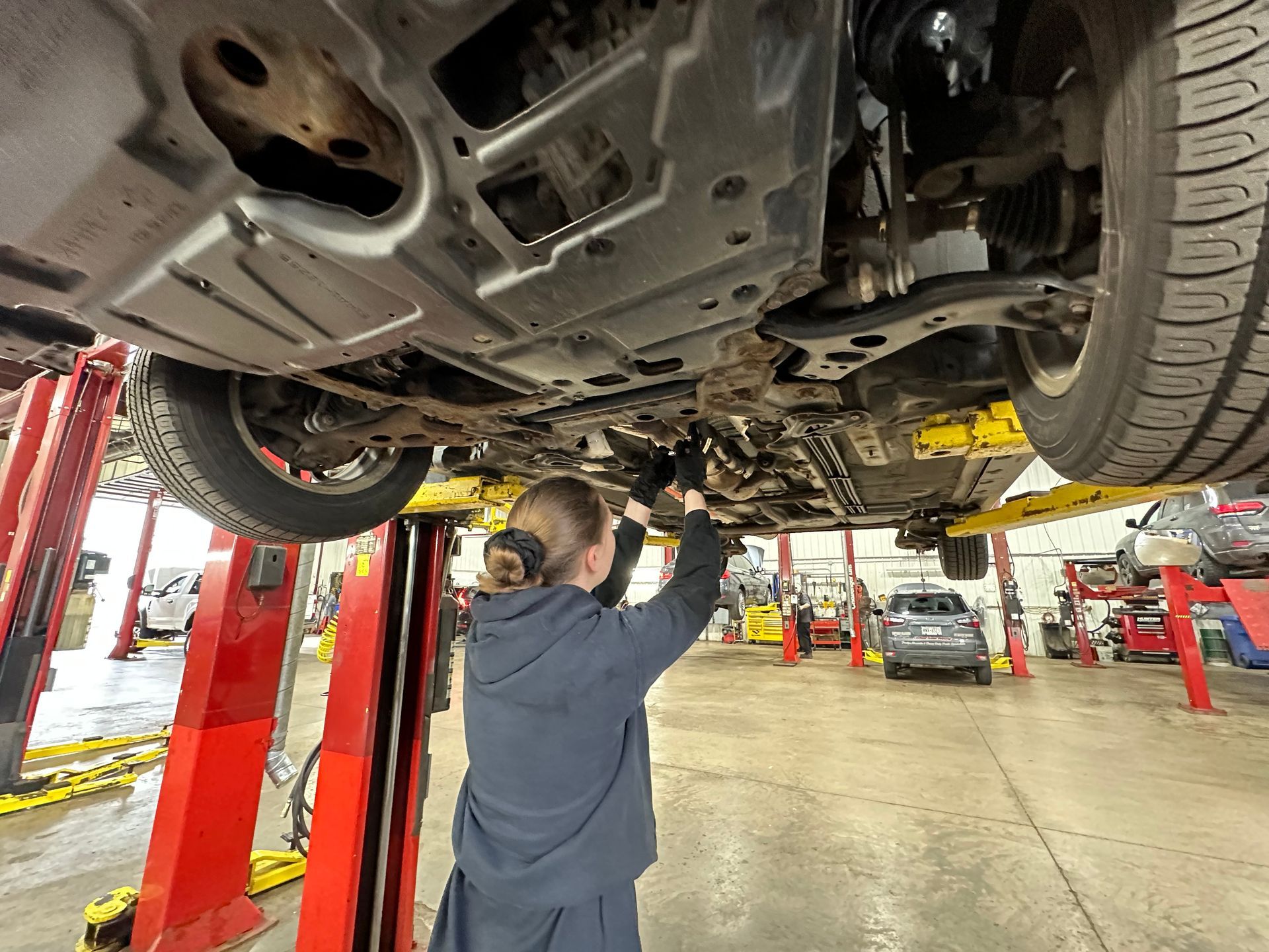 A woman is working under a car on a lift in a garage.  | Lou's Car Care Center, Inc.