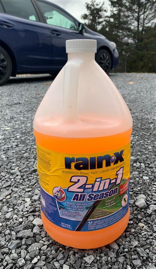 A gallon of rain x 2 in 1 all season sitting on gravel in front of a blue car.  | Lou's Car Care Center Inc.