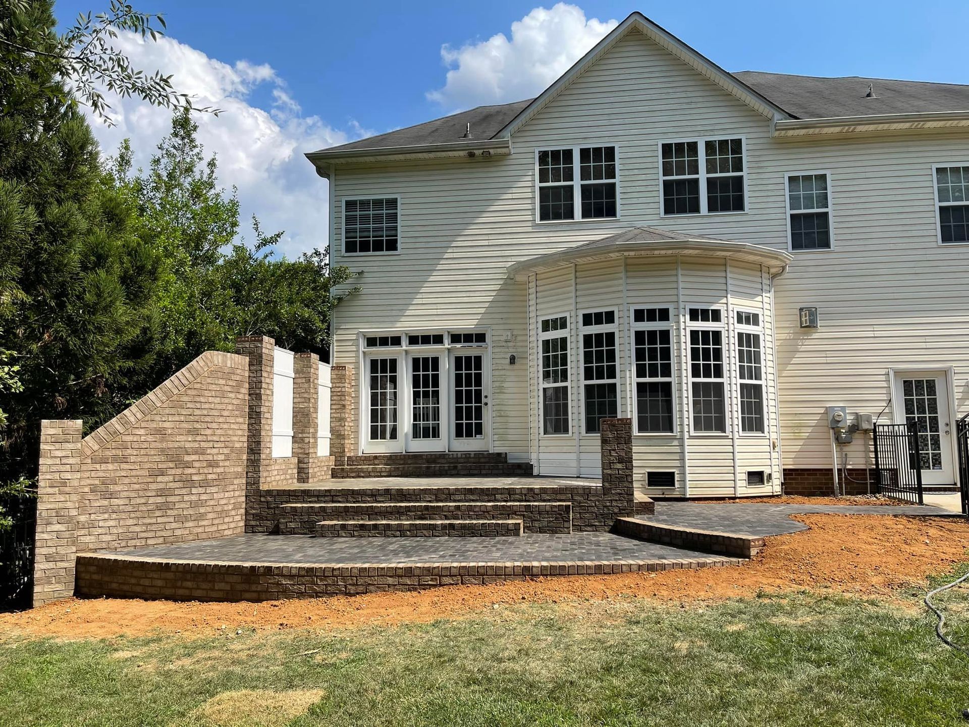 Back of a two-story beige house with a brick patio and steps leading to a yard, on a sunny day.