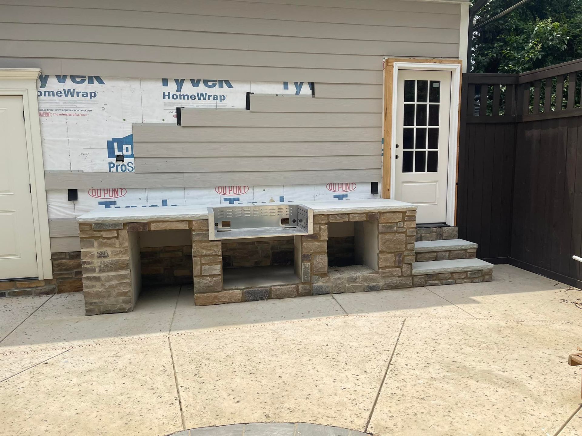 Outdoor kitchen with stone base and grill, next to a building with siding, on a concrete patio.