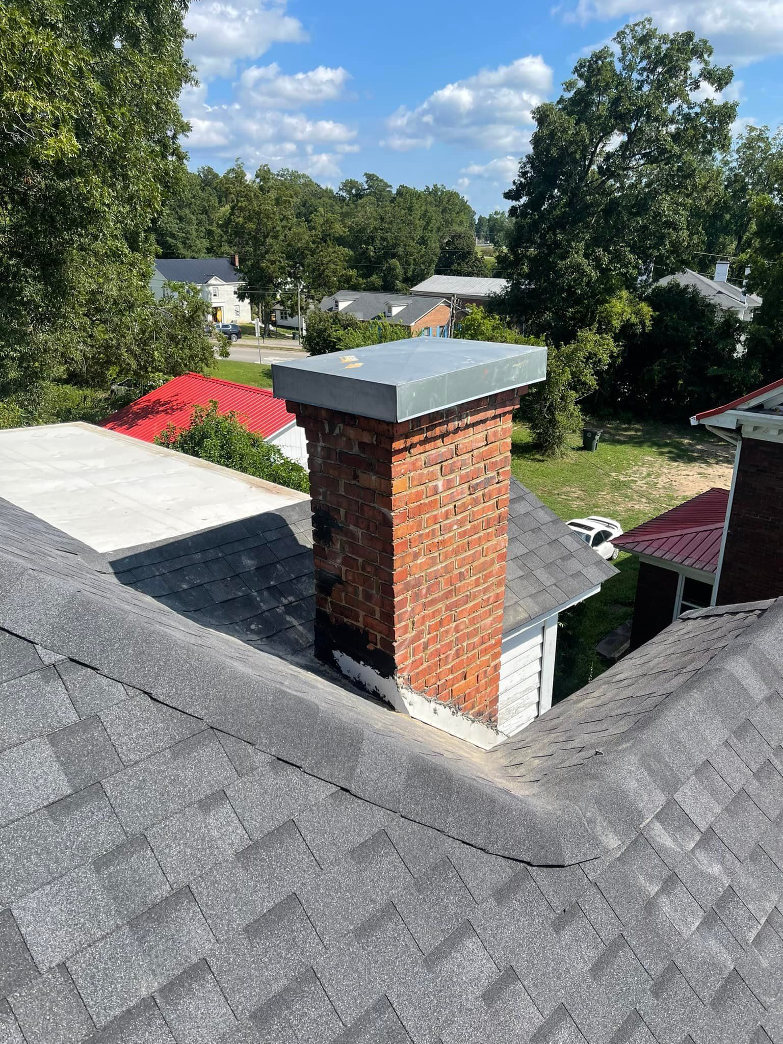 Brick chimney on a gray shingle roof, topped with a metal cap, under a blue sky.