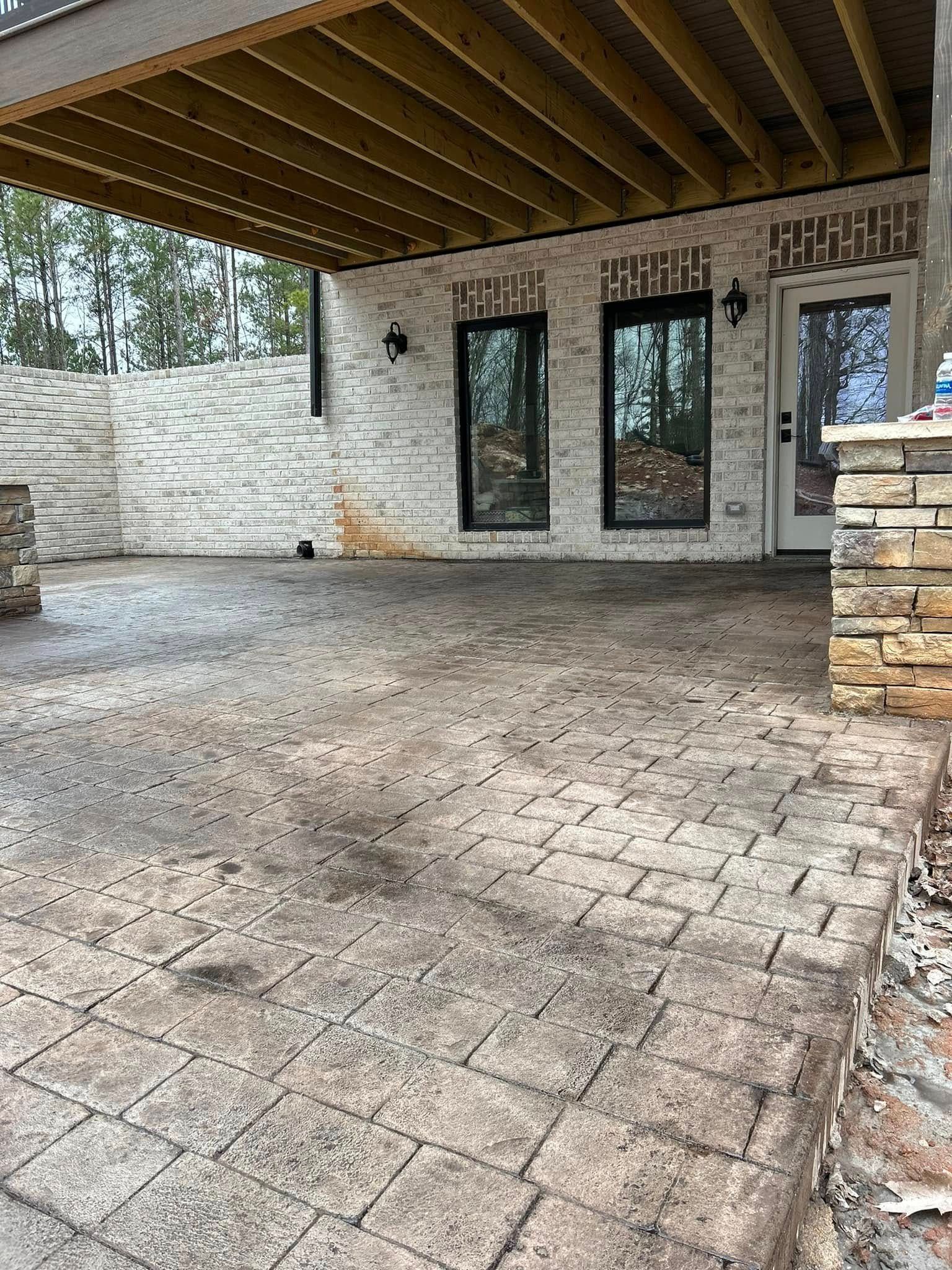 Patio with brick-like paving, beneath a wooden-beamed overhang. Features include brick walls and dark-framed doors.