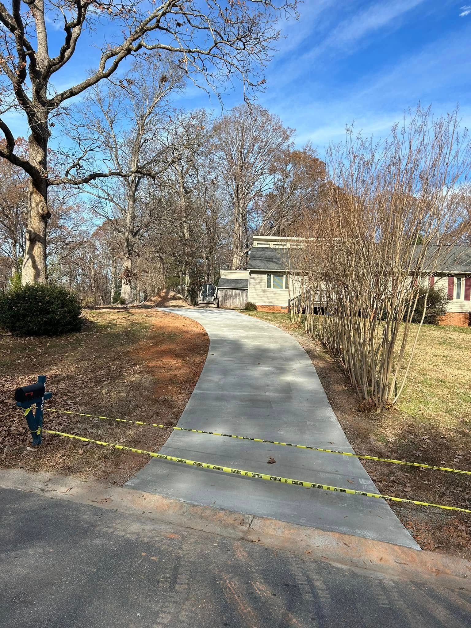 A concrete driveway bordered by yellow tape leads to a house with fall foliage on a sunny day.