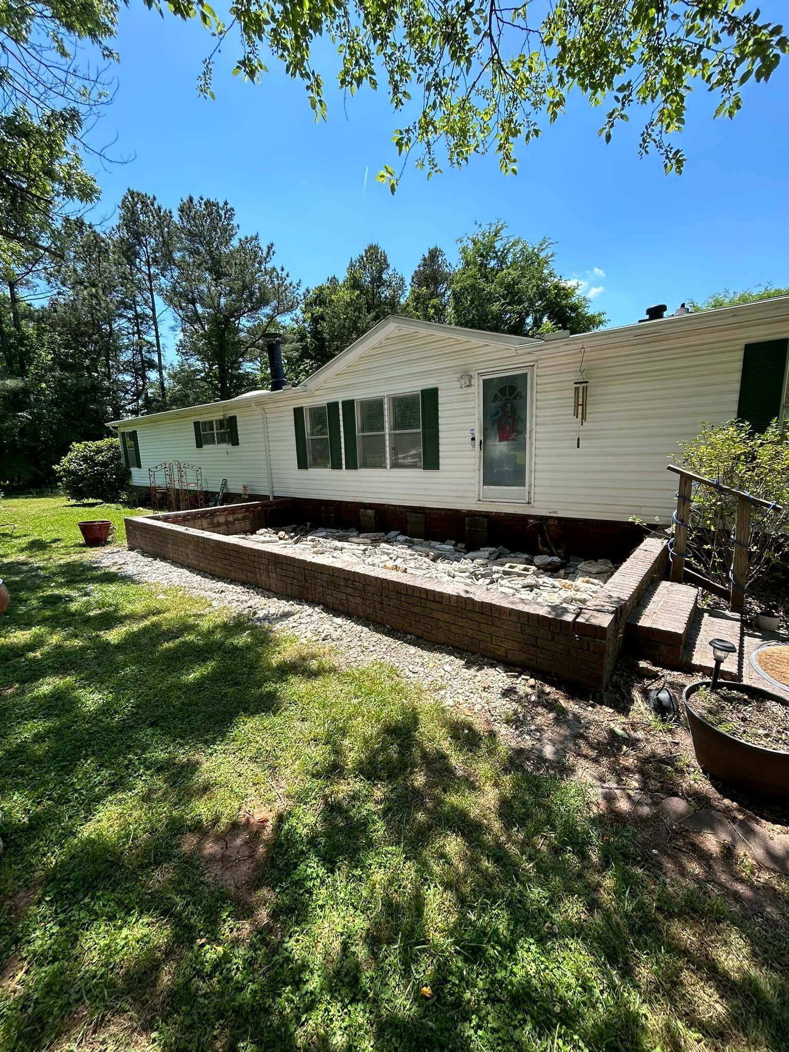 White house with green shutters, surrounded by grass and trees. A brick and gravel area in the foreground.