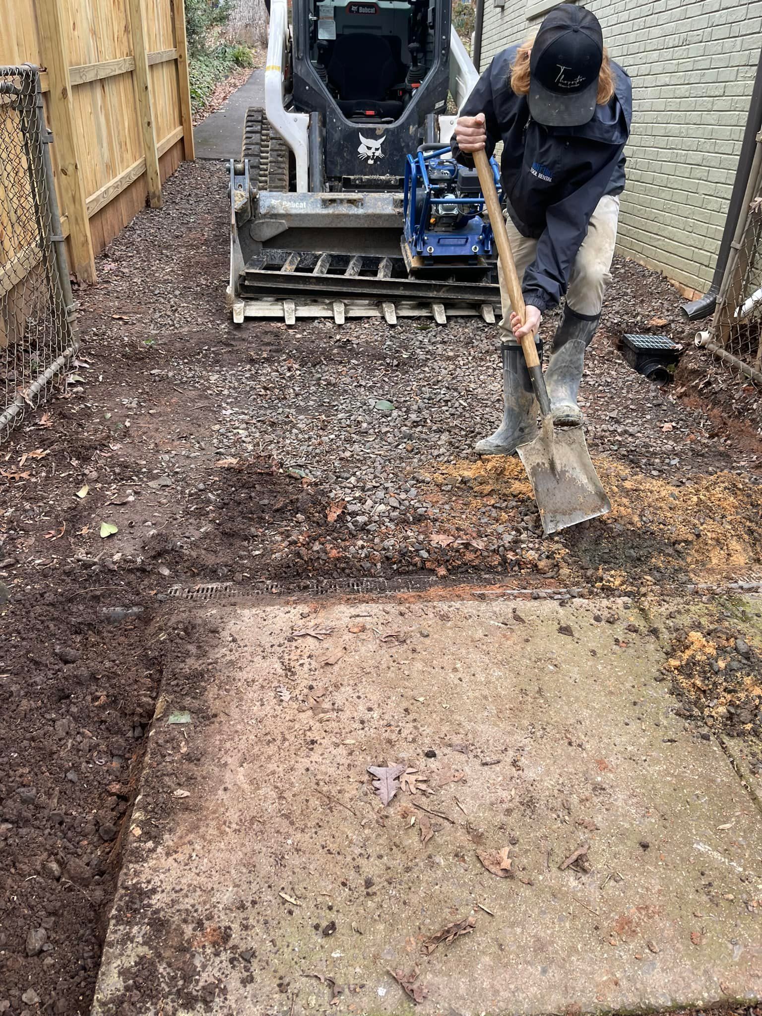 Person shovels soil near a Bobcat excavator and a concrete slab in a wooded area.