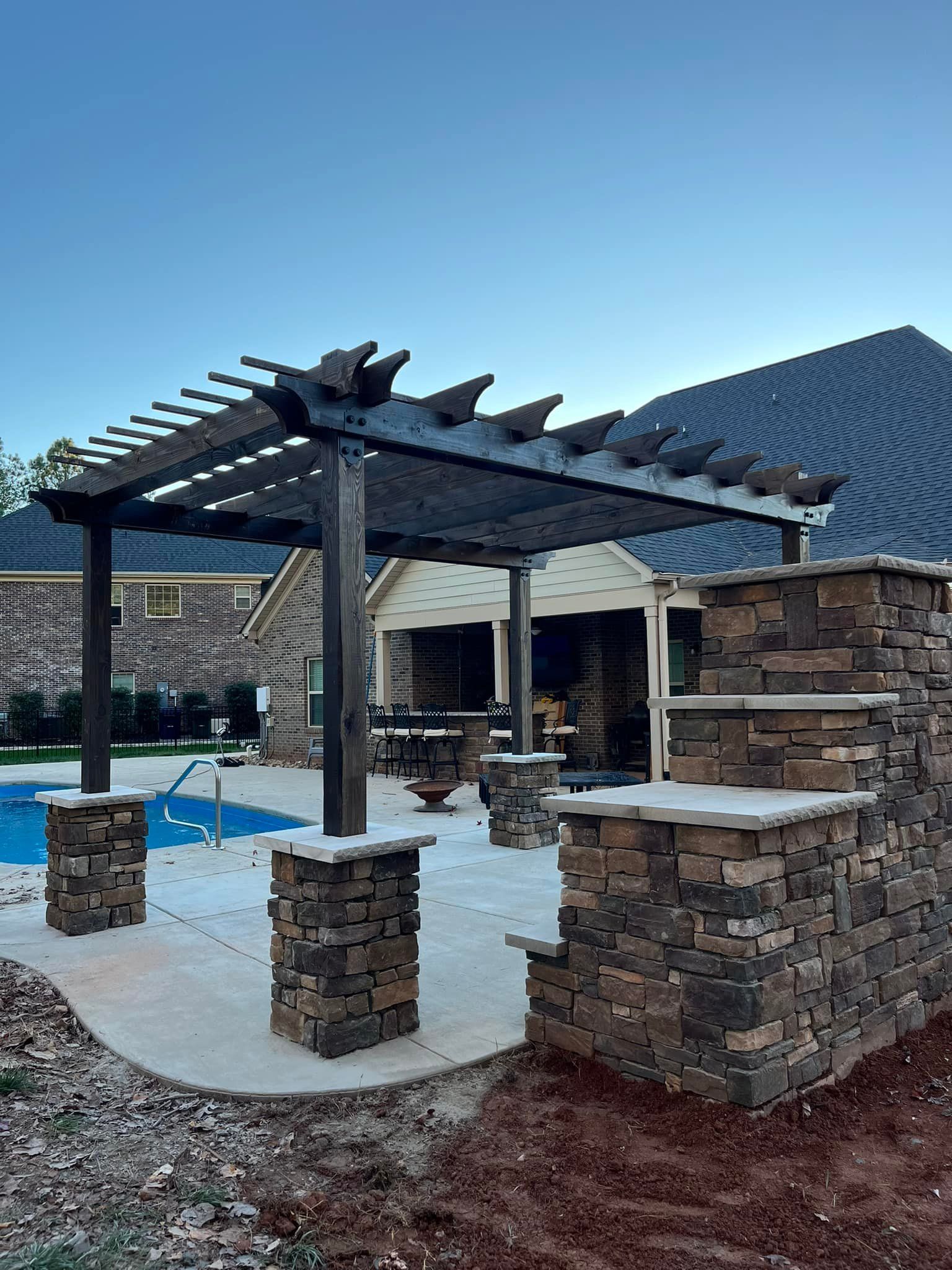 Pergola with stone columns over a patio, next to a pool and a house with a dark roof.
