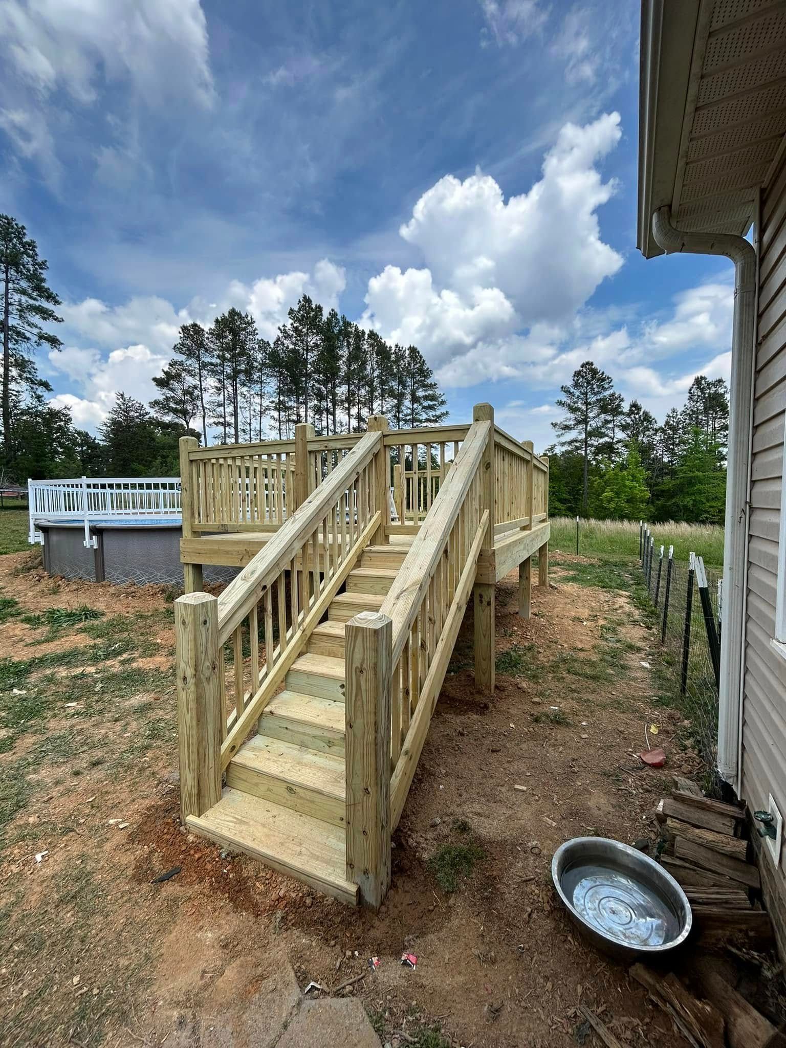 Wooden deck with stairs, built next to a house. An above-ground pool sits in the background, against a cloudy sky.