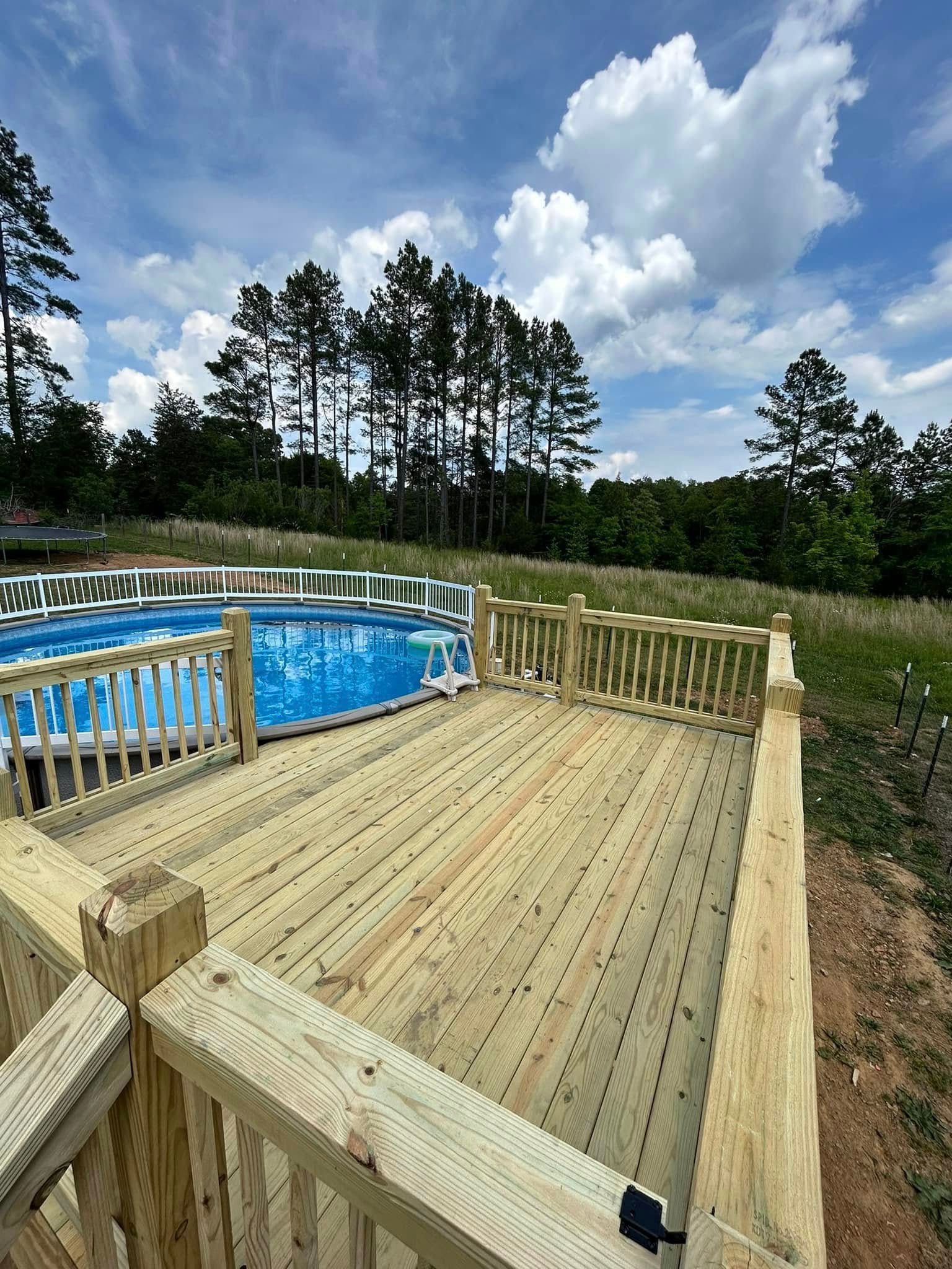 Wooden deck with a blue above-ground pool, against a background of trees and a cloudy sky.