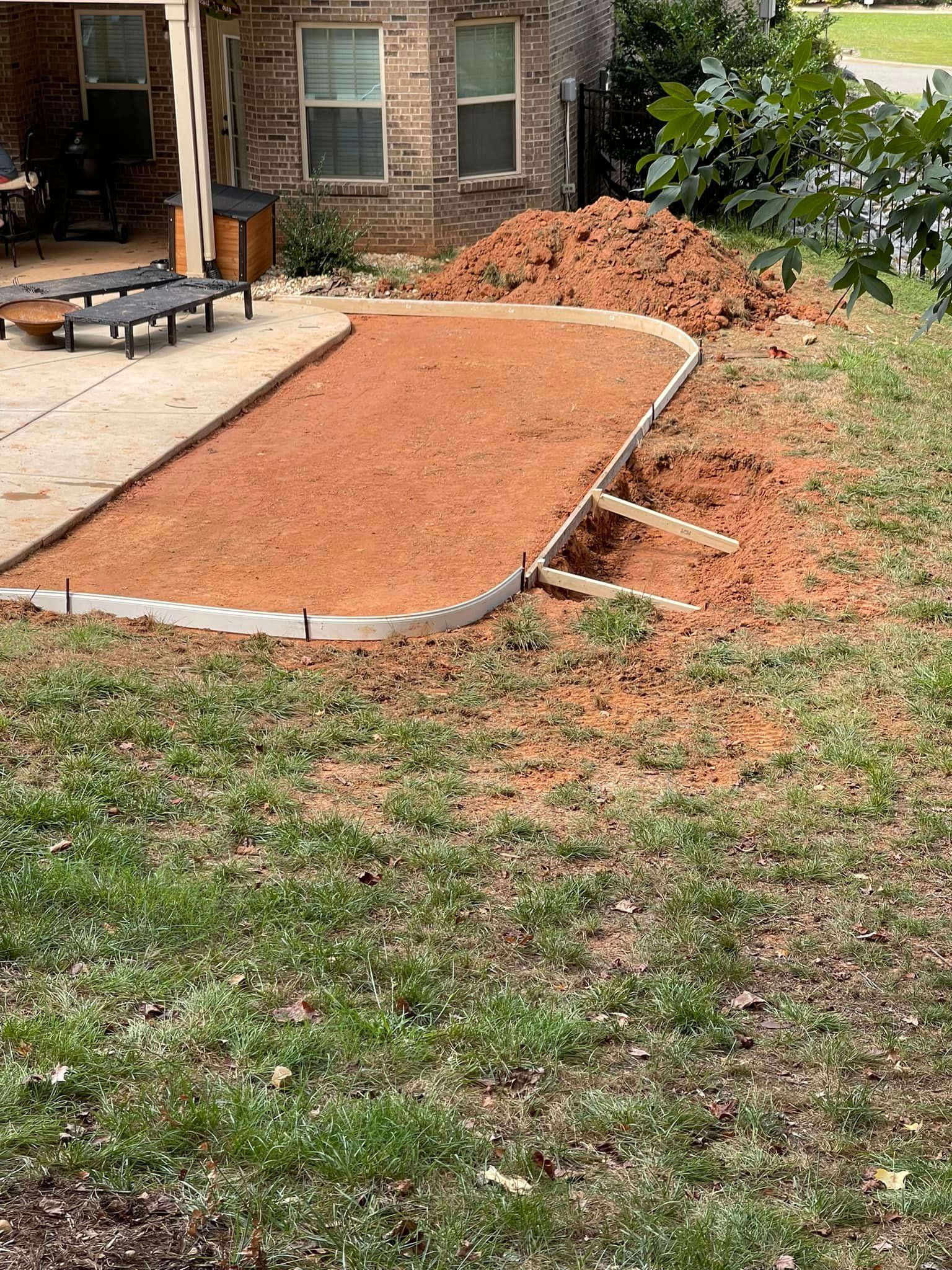 Construction site: concrete border framing a dirt-filled area beside a patio; pile of dirt and grass surround.