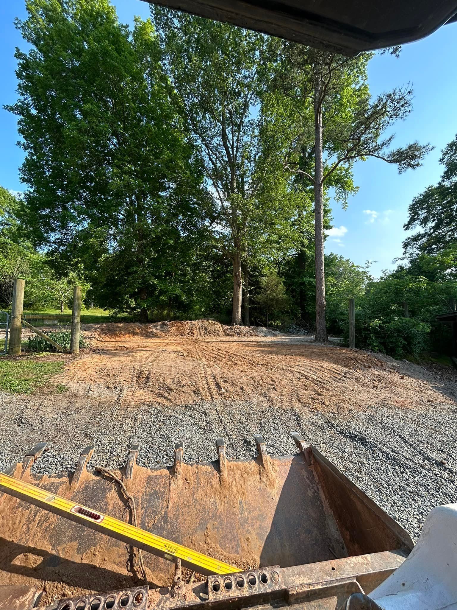 A construction site with gravel, dirt, trees, and an equipment bucket. The sky is visible.