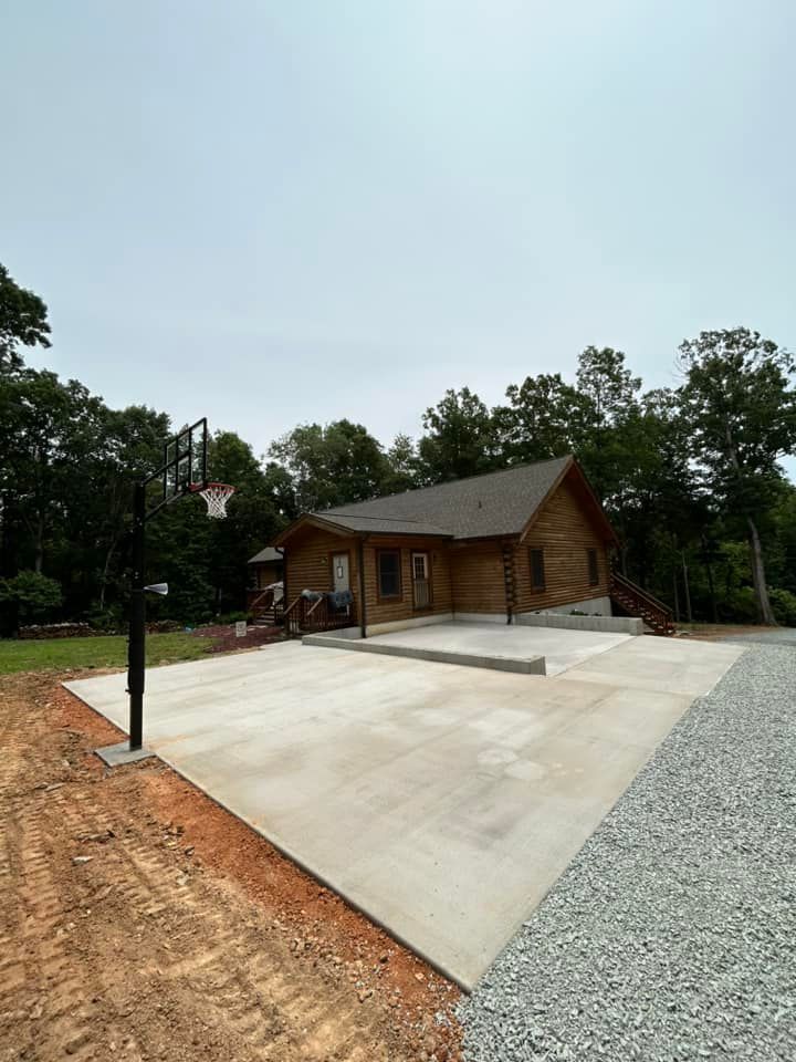 Concrete basketball court with hoop, next to a log cabin, surrounded by trees. Cloudy sky.