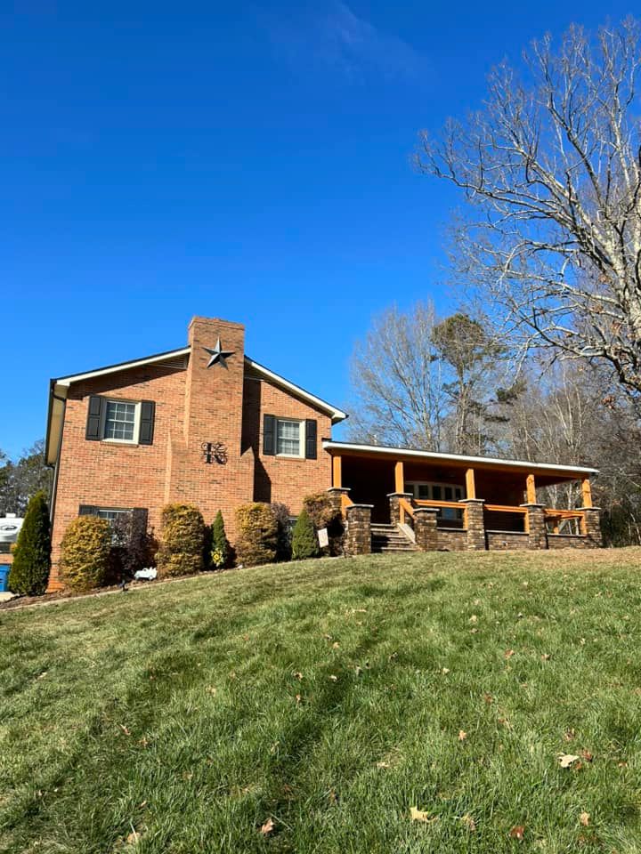 Brick house with porch and chimney. Green lawn, blue sky, and a bare tree are in the background.
