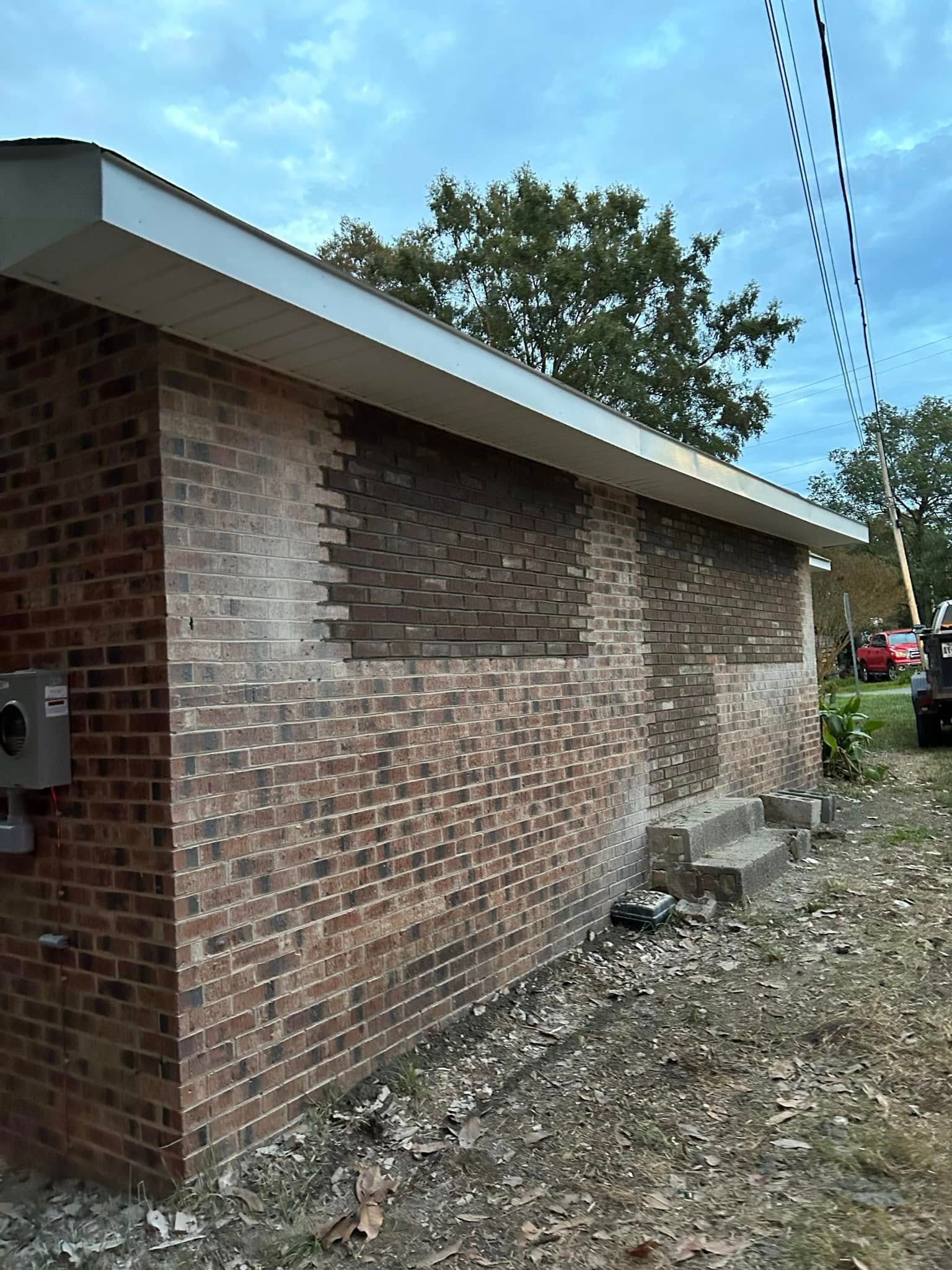 Brick building exterior with patched-over window spaces. Electrical box on left. Steps on right. Green foliage and power lines in the background.