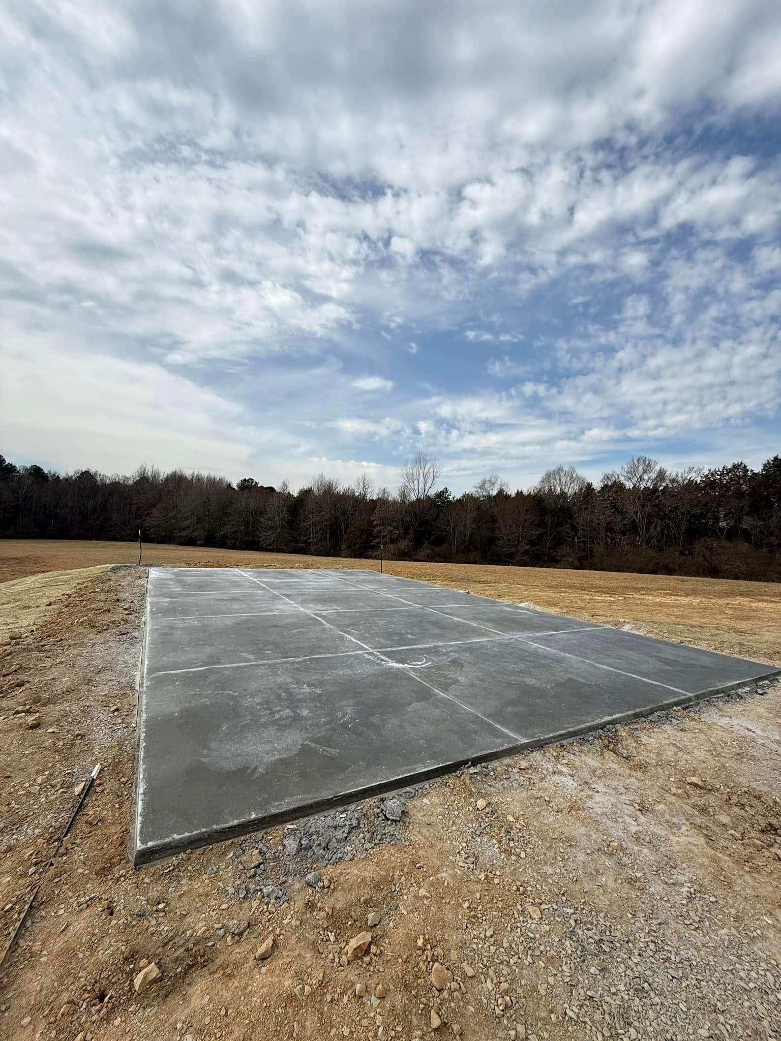 A rectangular concrete foundation on a dirt lot, under a cloudy blue sky. Trees in the background.