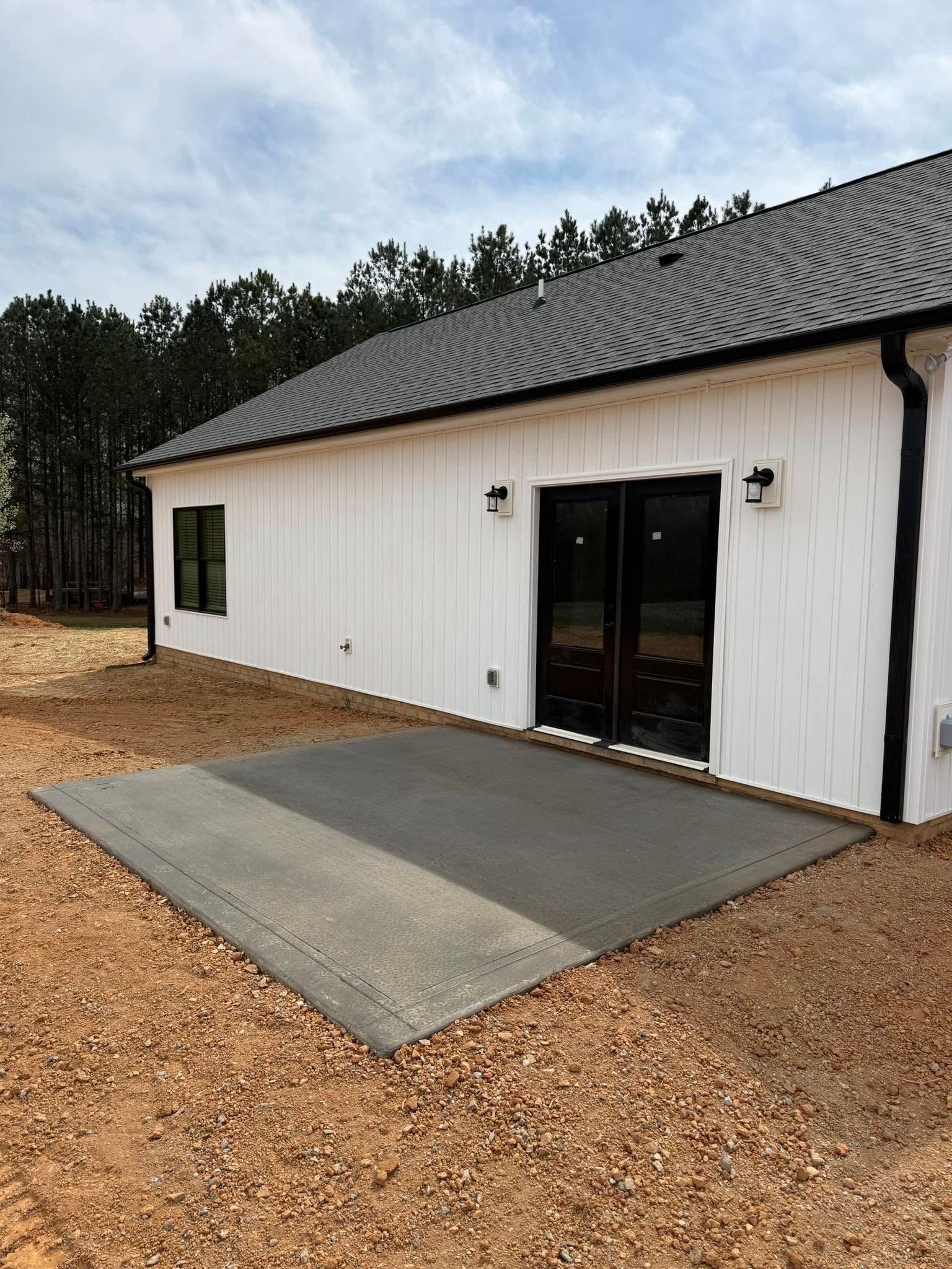 Exterior of a white house with a concrete patio. Black trim, two sconces, and sliding glass doors. Brown gravel ground.