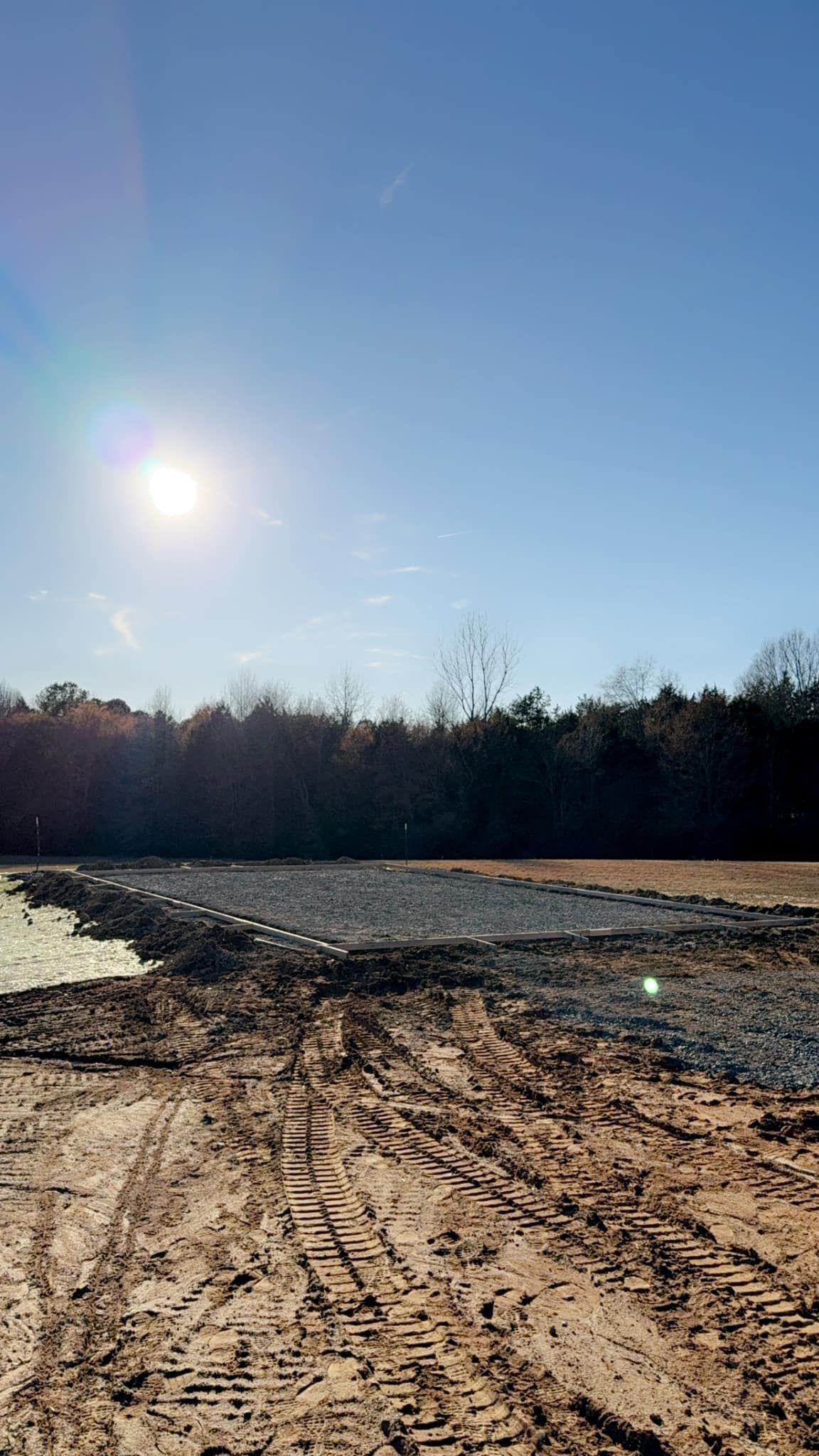 Sunny day over construction site with mud, tire tracks, and a treeline.