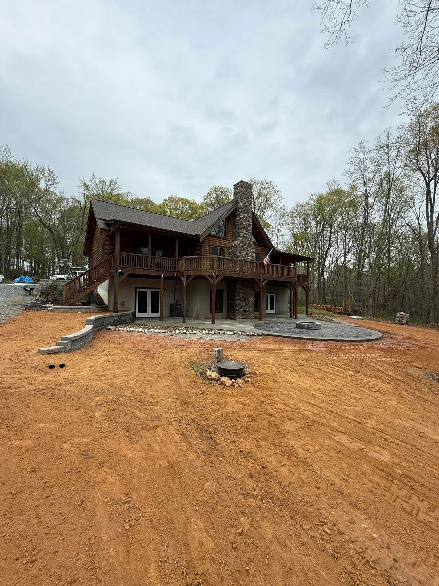 Log cabin with stone chimney and wrap-around deck on a hillside with freshly turned earth.
