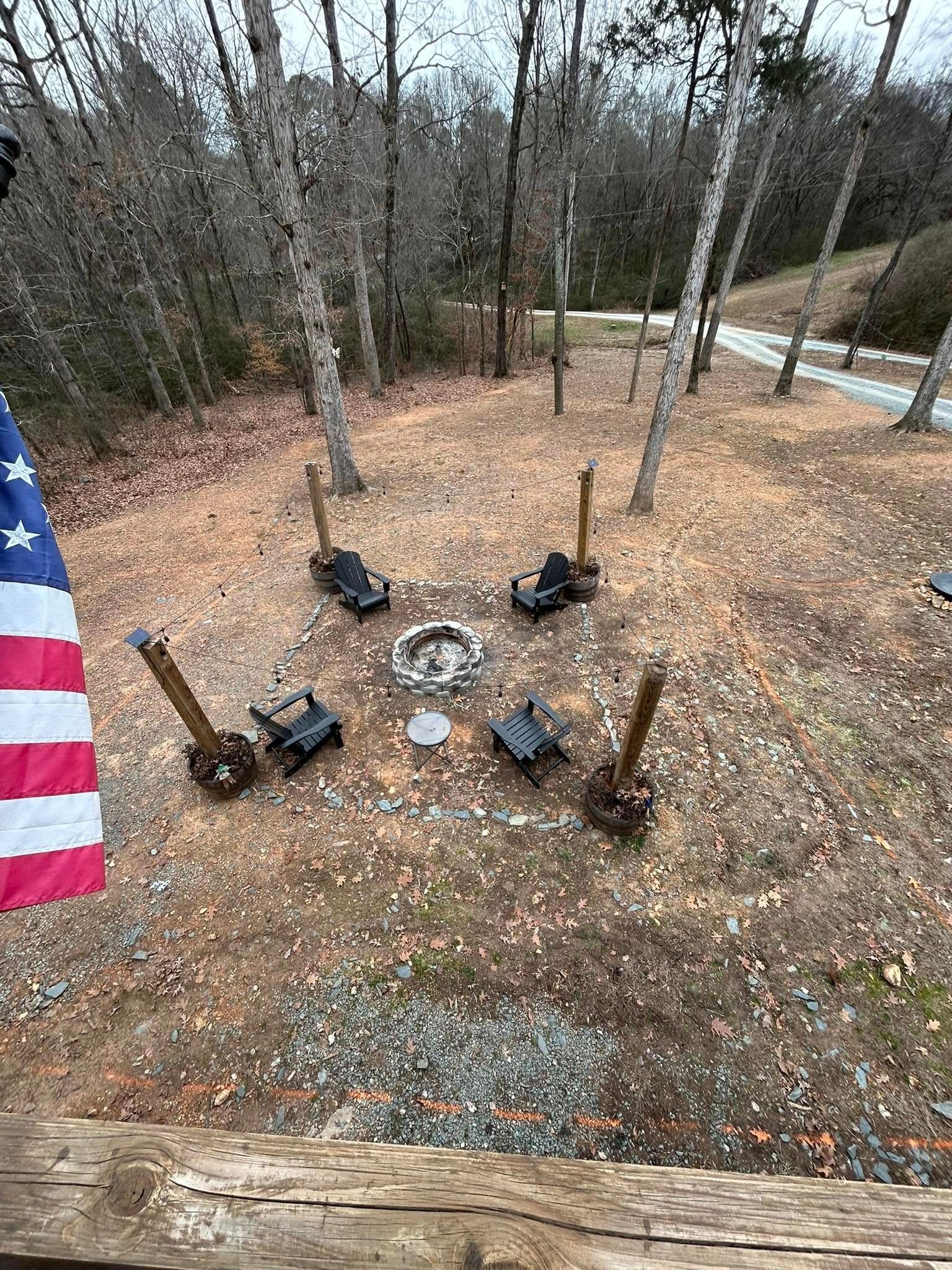 Fire pit surrounded by chairs in a wooded area, with an American flag to the left.