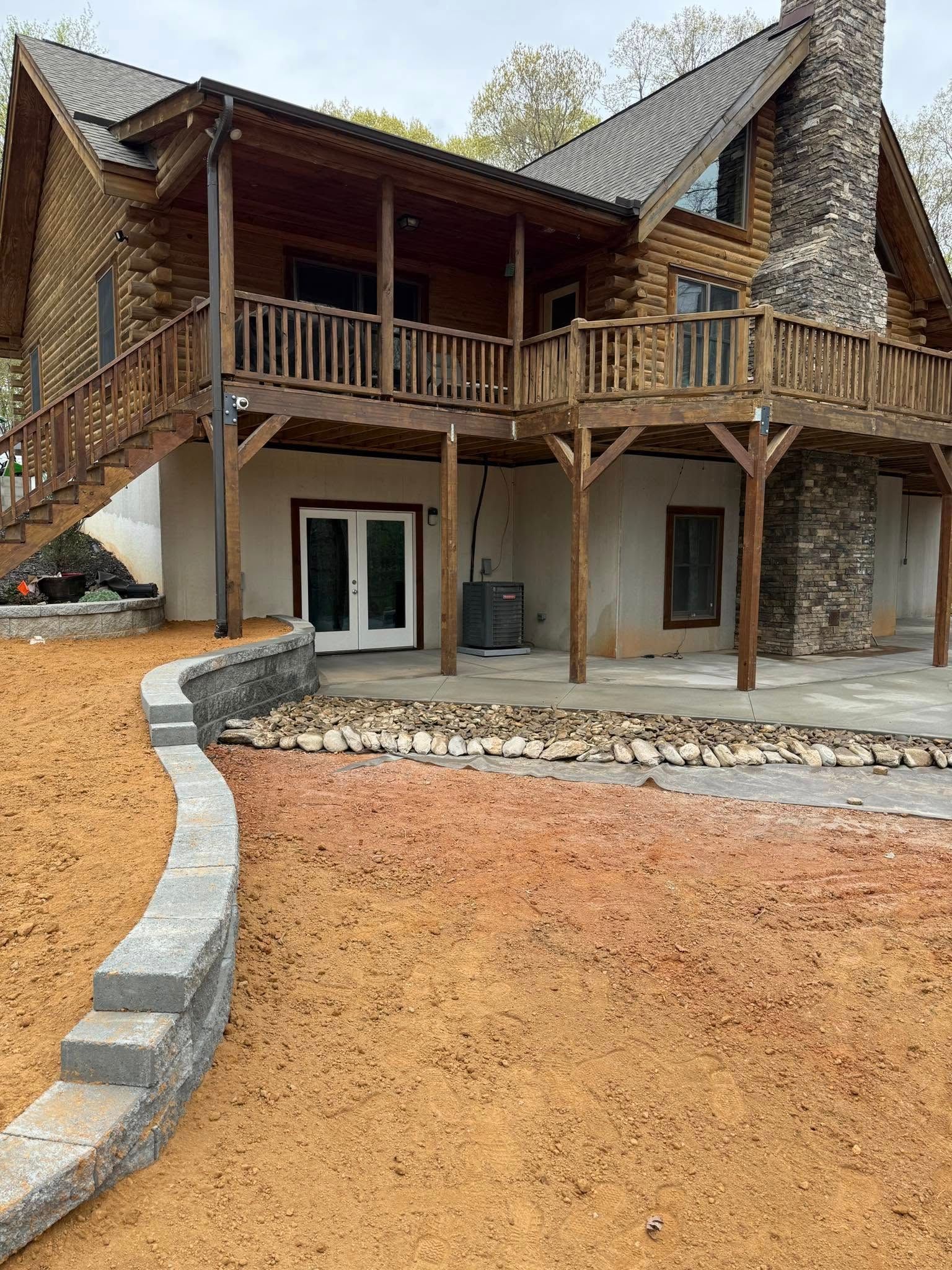Log cabin with stone chimney, wooden deck, and retaining wall. Exterior view of the house on a bright day.
