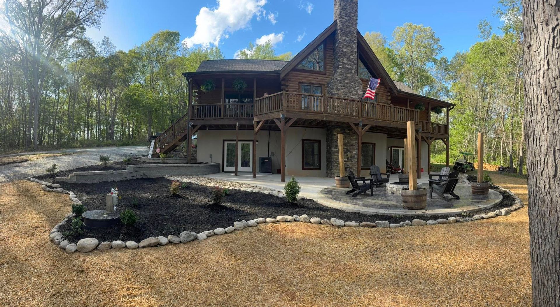 Cabin with stone chimney and large deck, landscaped yard with fire pit and gravel pathway.
