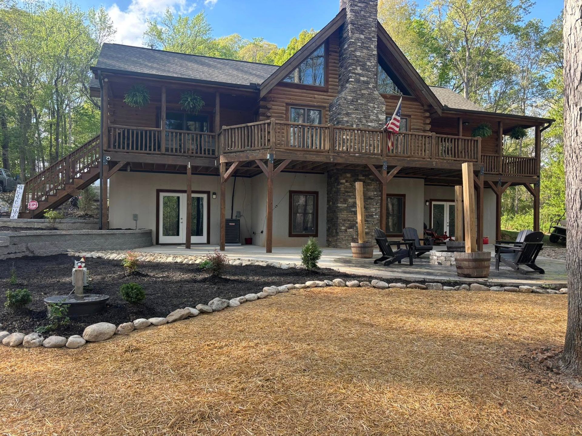 Wooden cabin with a large deck, stone chimney, and landscaping in a wooded setting.