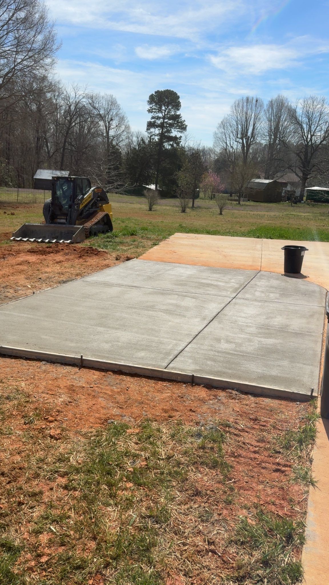 Concrete slab construction site with skid steer, grassy ground, trees, and blue sky.