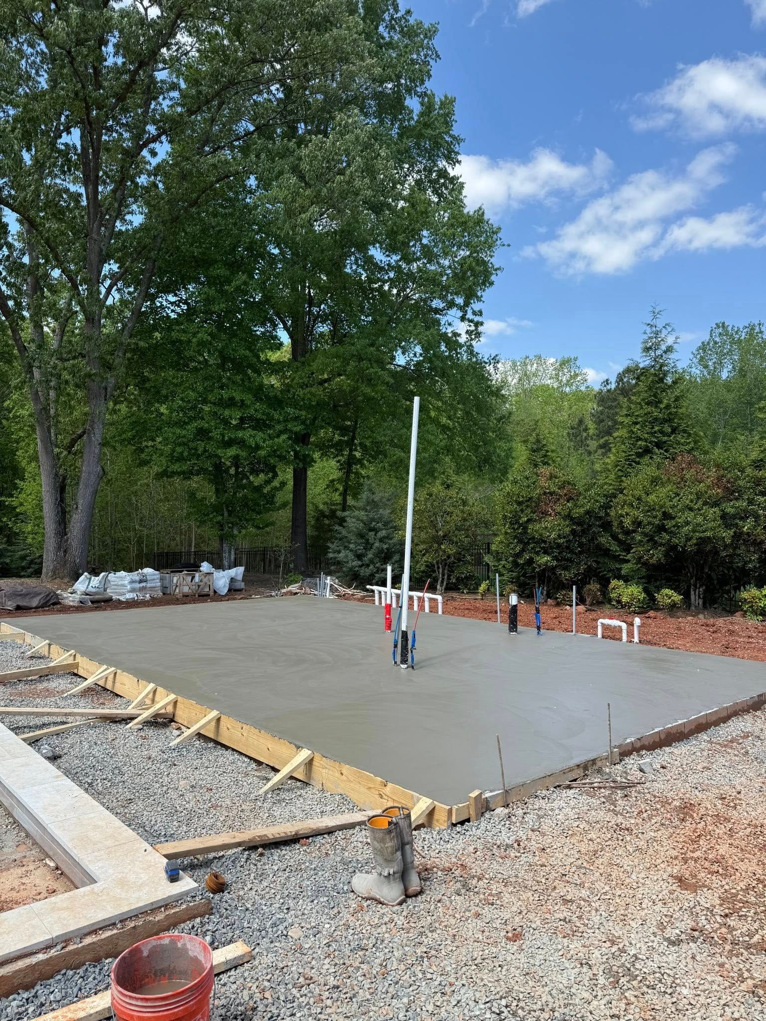 Freshly poured concrete foundation with plumbing pipes, surrounded by gravel and wooden forms, under a blue sky.