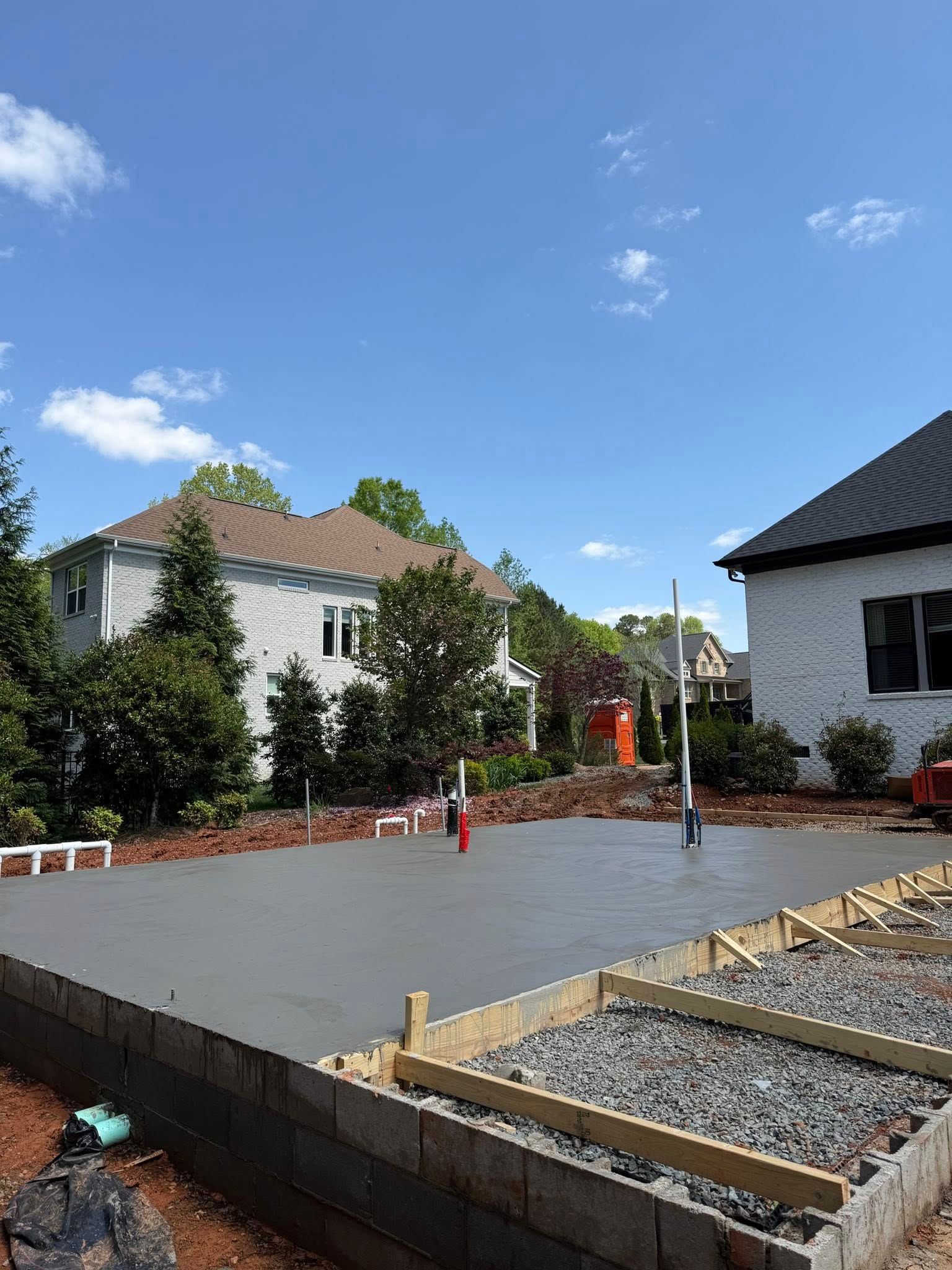 Newly poured concrete foundation with plumbing, surrounded by wooden forms, in front of houses under a blue sky.