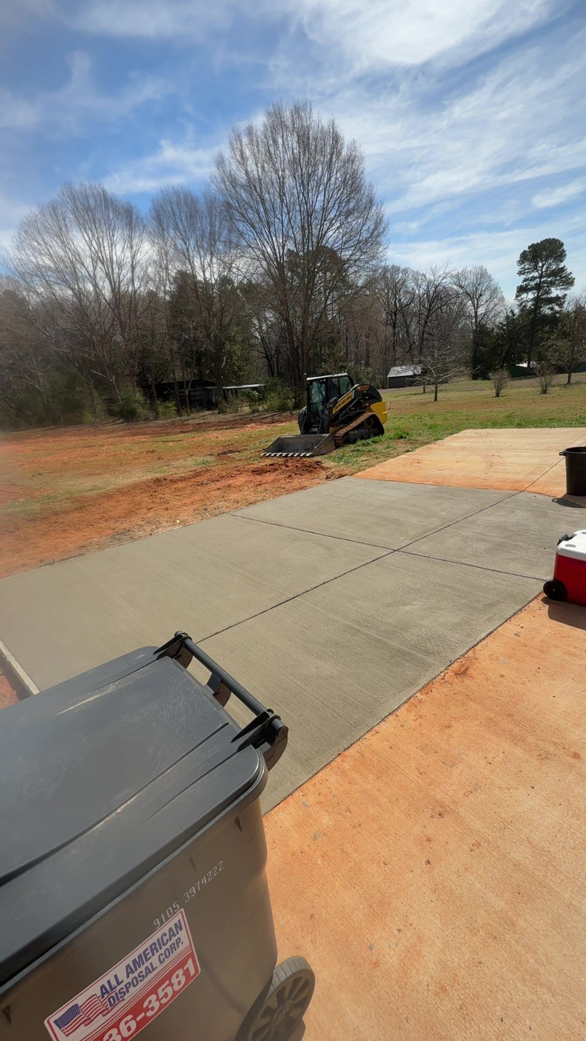 Skid steer operating in a yard; newly poured concrete driveway in foreground; sunny, blue sky.