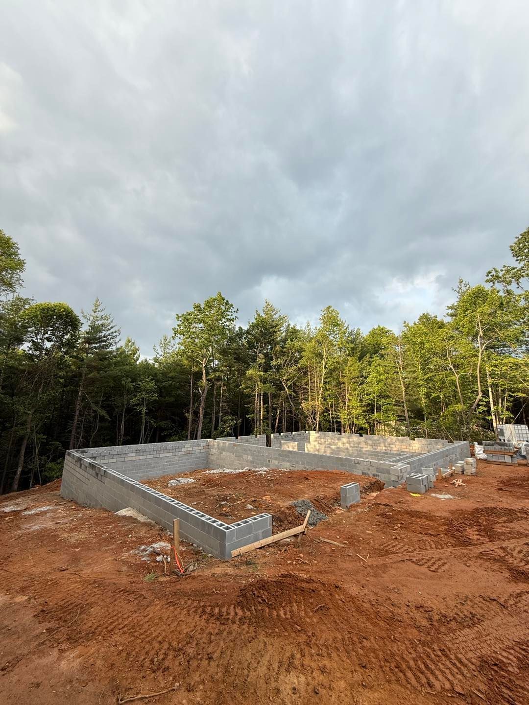 Foundation of a building constructed with concrete blocks on a dirt lot, surrounded by trees under a cloudy sky.