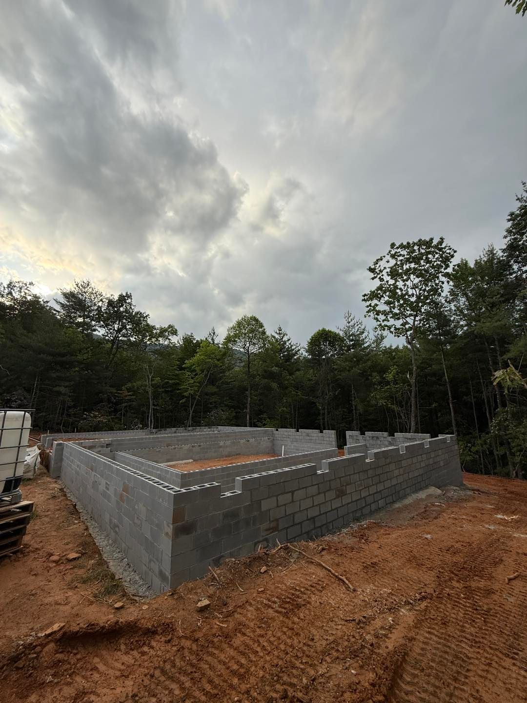 Foundation of a building constructed with gray cinder blocks on a dirt lot, overcast sky, trees in the background.