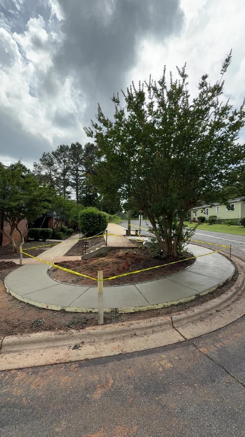 New concrete sidewalk curves around a tree and garden bed on a cloudy day.