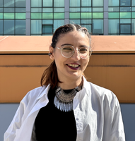 A woman wearing glasses and a necklace is smiling in front of a building.