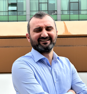 A man with a beard is smiling with his arms crossed in front of a building